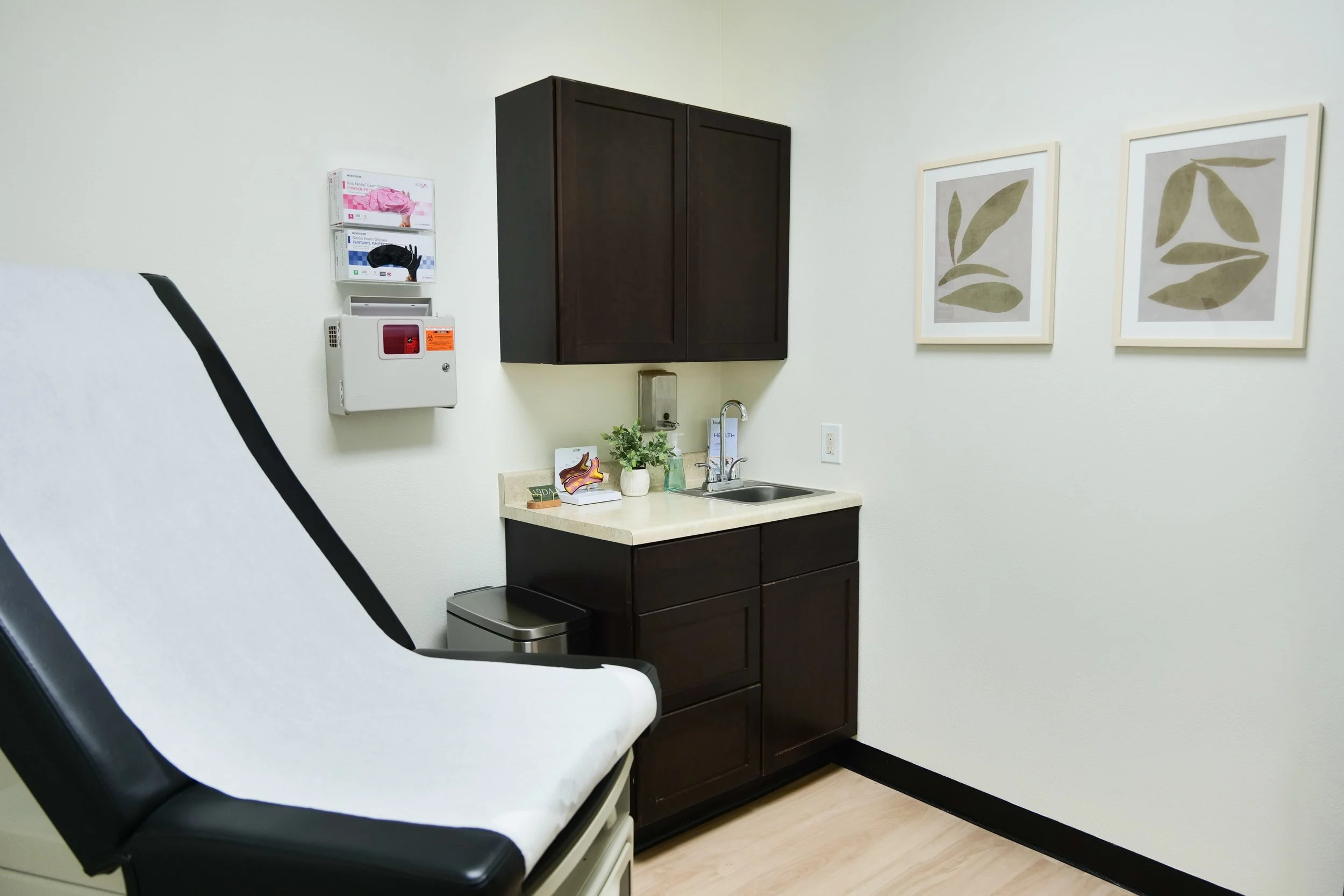 Medical examination room with an examination table, dark wooden cabinets, framed leaf art on the wall, and a small sink with a plant and medical pamphlets on the counter.