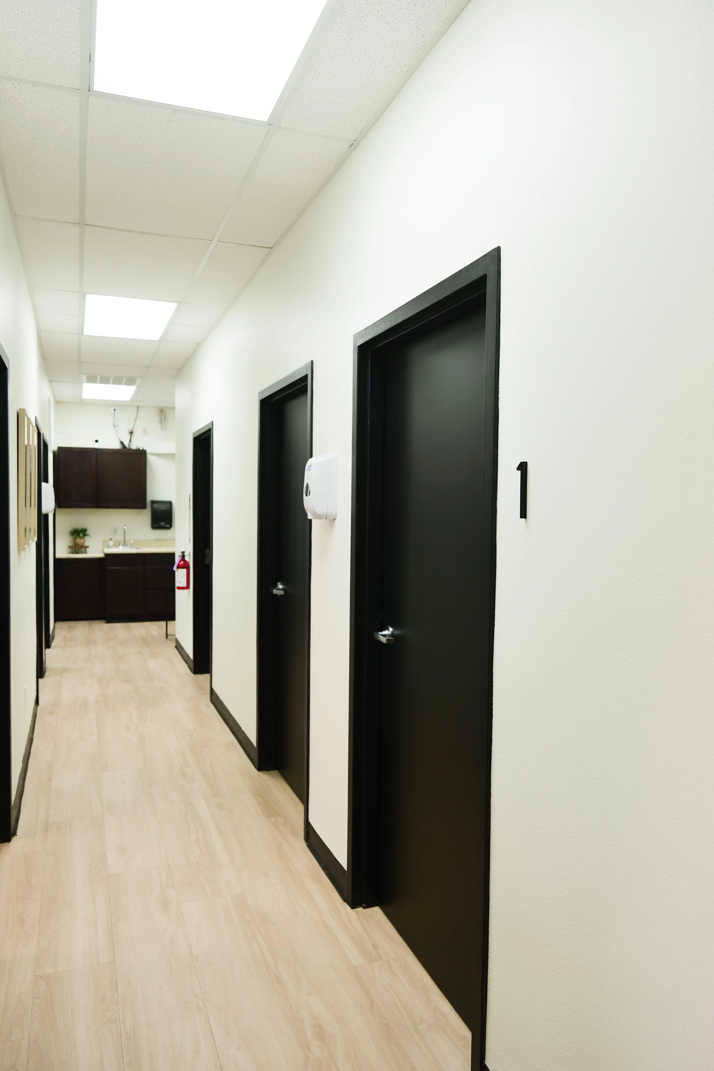 Medical Clinic Hallway with white walls, black doors with numbers, and light wood floor.