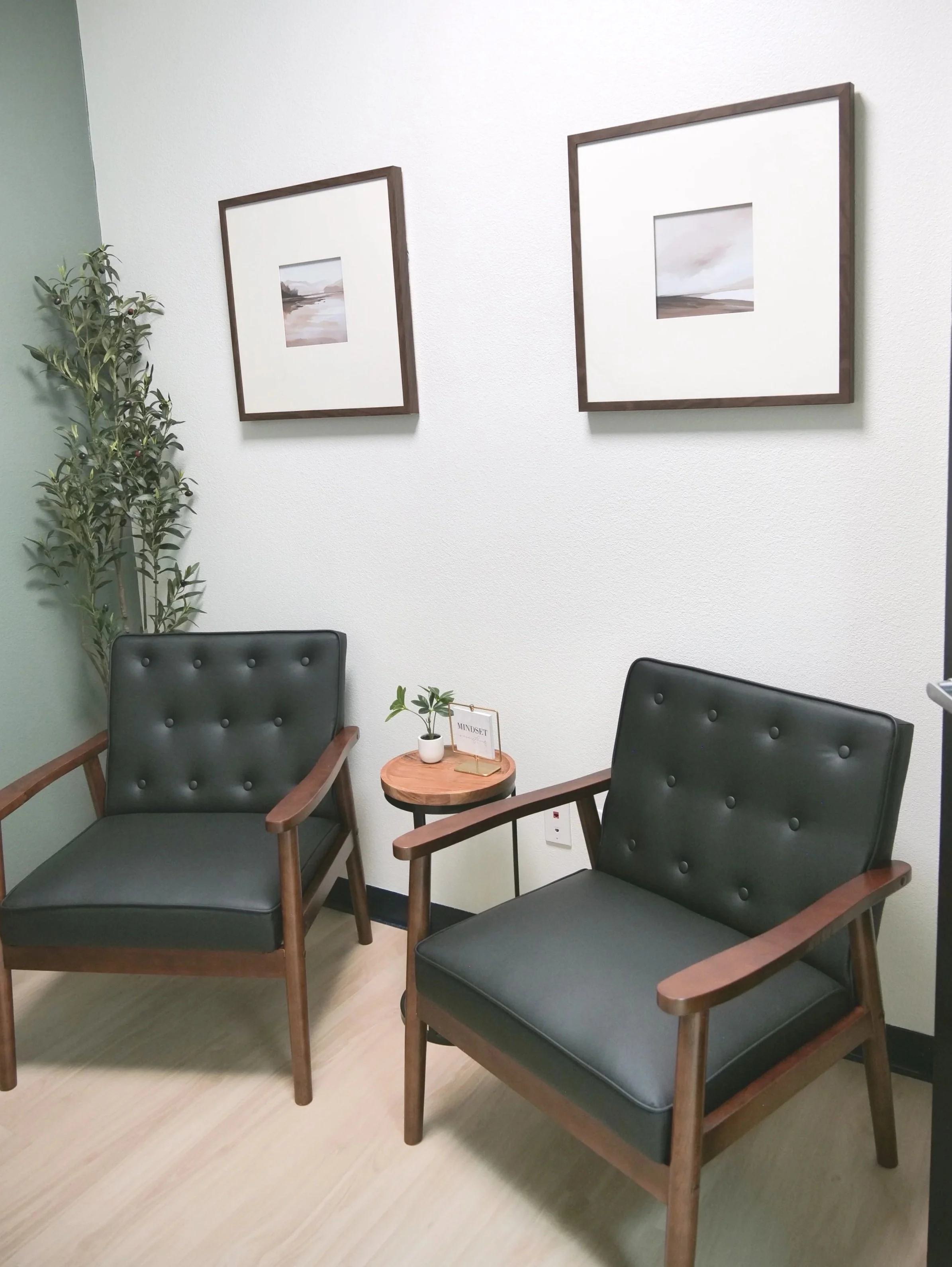 Two black armchairs with wooden frames, a small round wooden side table with a small plant and a paper sign, potted plants in the corner, and two framed landscape paintings on a white wall in a waiting room.