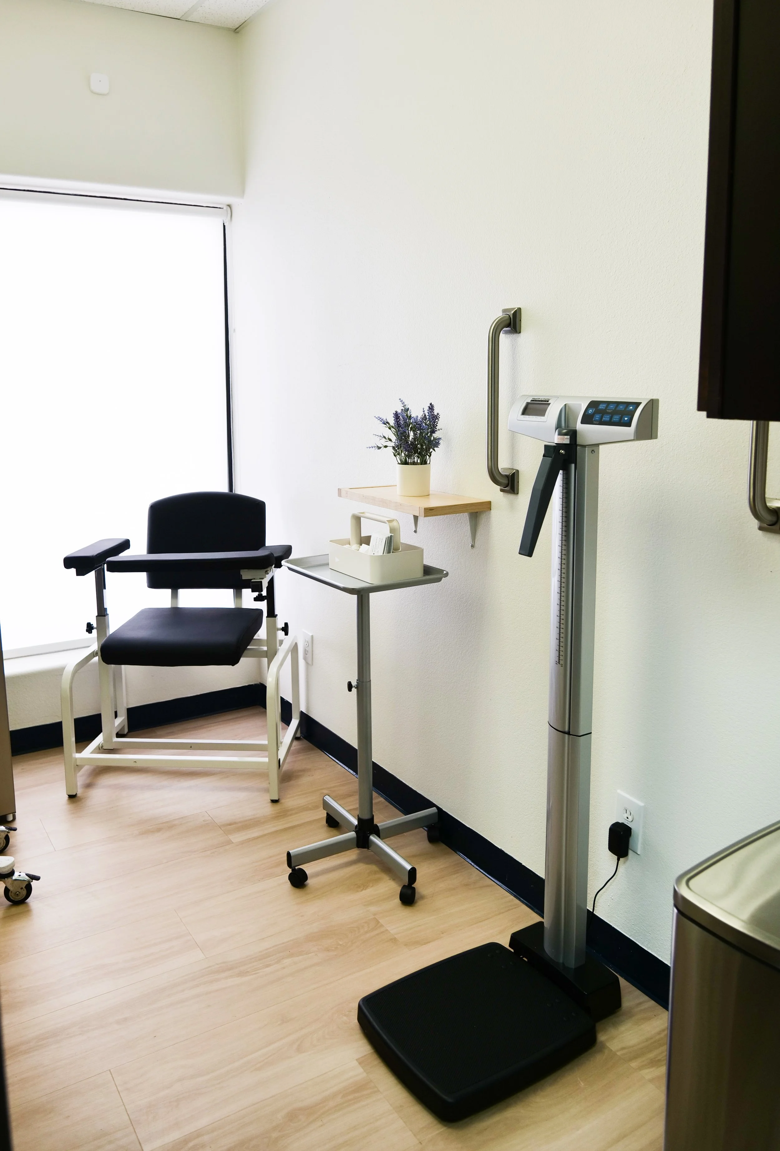 Medical examination room with a wheelchair, a foot scale, a small table with a potted plant, and medical equipment. Bright lighting and hardwood floors.
