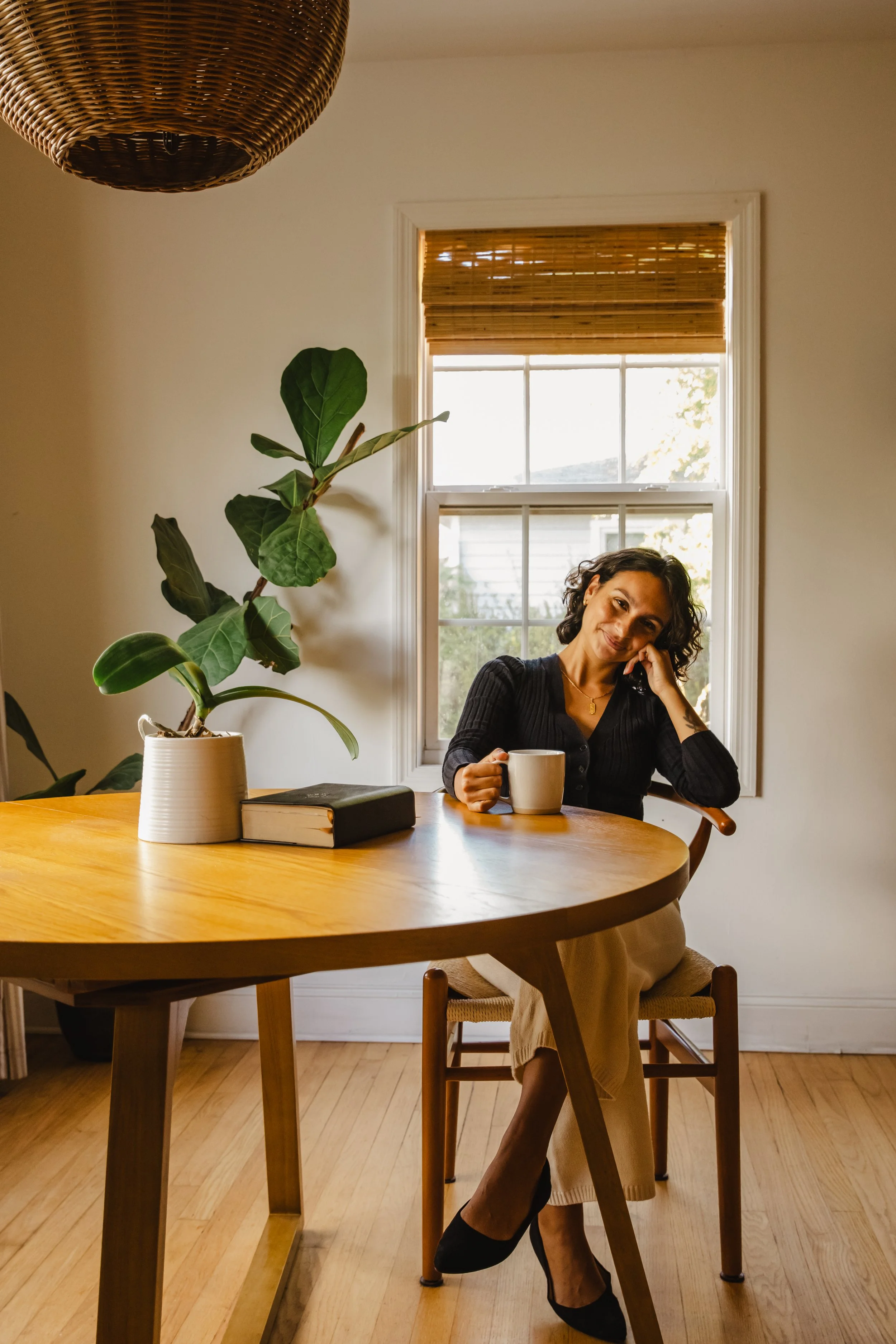 A woman sitting at a wooden dining table holding a white mug and smiling, with a large green plant on the table and a window with a bamboo shade behind her.