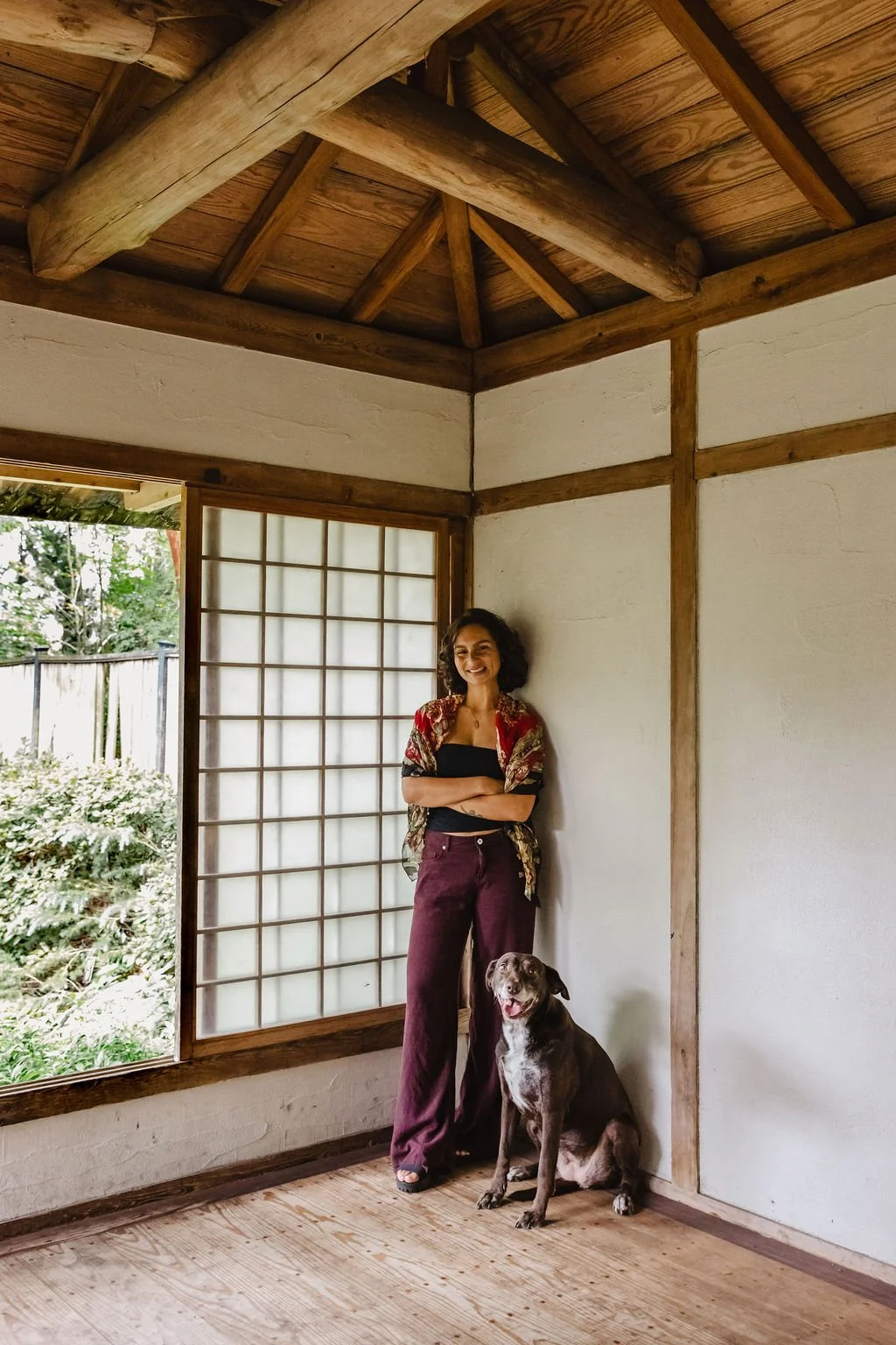 A woman and a dog inside a traditional Japanese-style room with shoji screens and wooden beams, with greenery visible outside the open window.