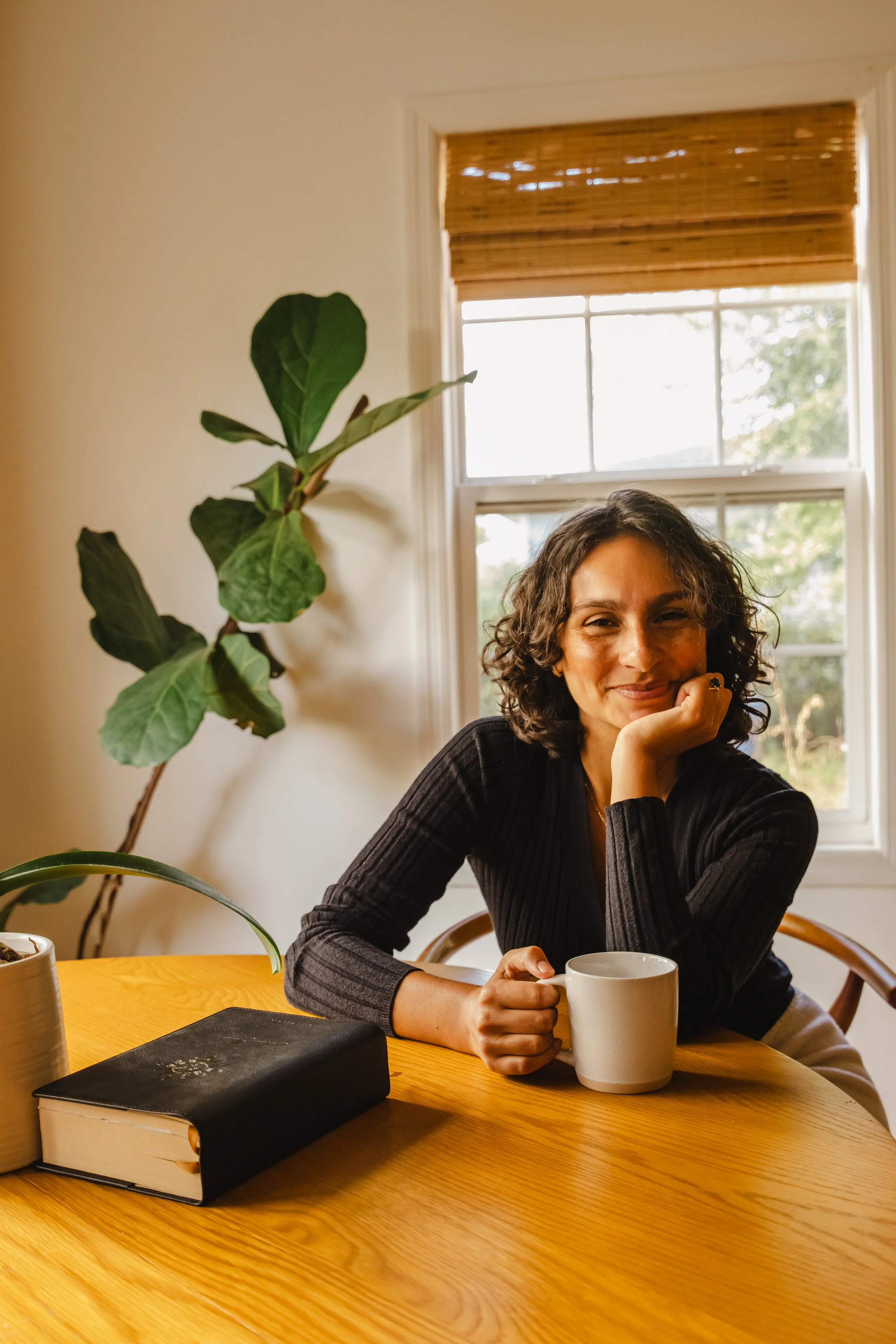 A woman with curly dark hair smiles while sitting at a wooden table, holding a white mug. The background features a window with bamboo blinds and a green plant next to her.