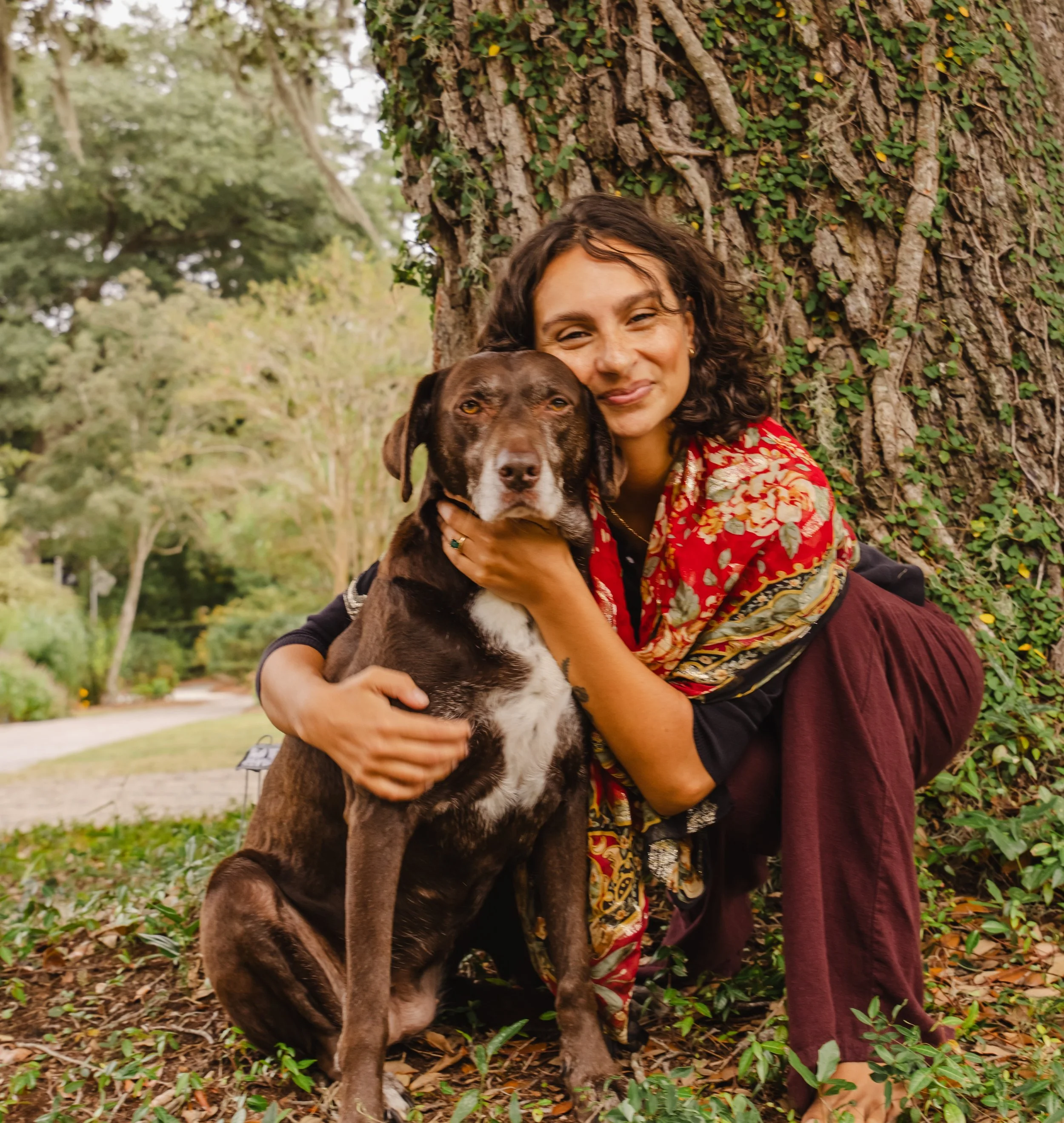 A woman hugging a brown and white dog outdoors near a large tree with green ivy and leaves.