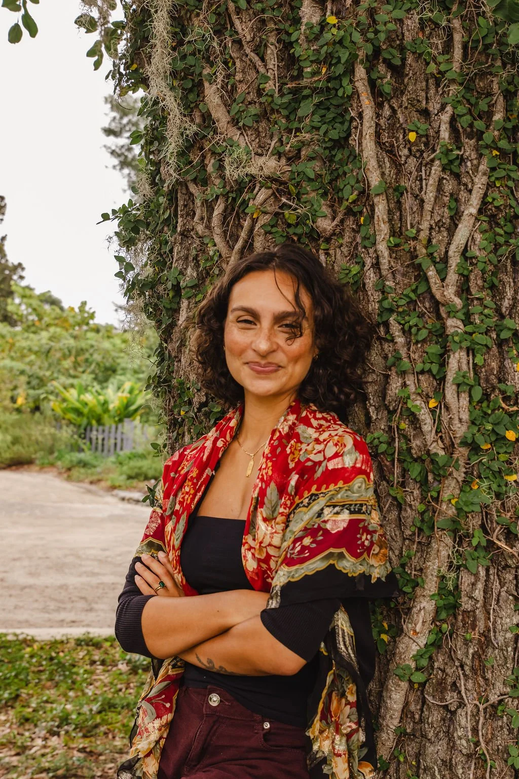 A woman with shoulder-length curly hair smiling and crossing her arms, standing outdoors next to a large tree with textured bark and green ivy.