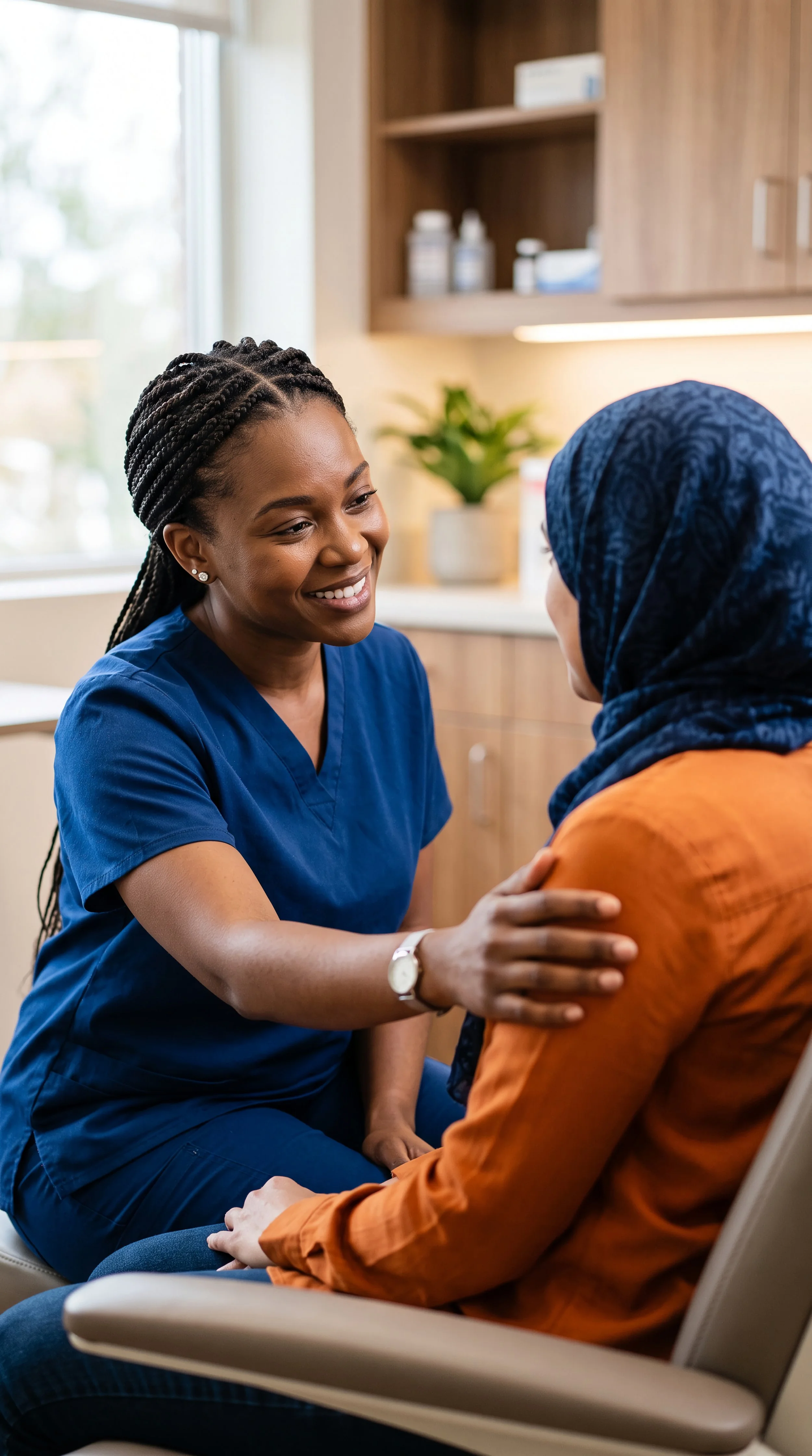 A healthcare professional, wearing blue scrubs, is speaking with a patient who is wearing an orange shirt and a blue headscarf. The healthcare worker is smiling warmly, with her hand on the patient's arm, in a medical office setting.