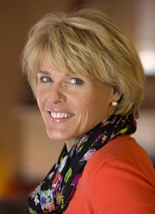 A woman with short blonde hair smiling, wearing a floral scarf, pearl earrings, and an orange top.