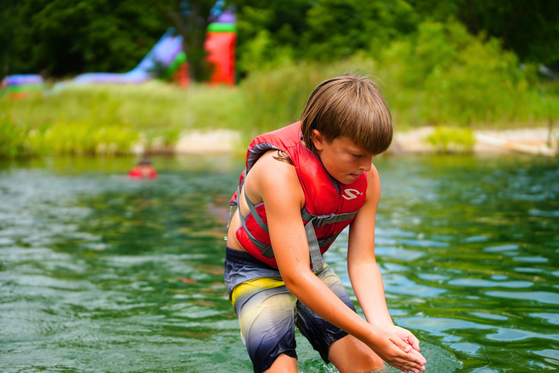 A young boy wearing a red life jacket and gray shorts standing in a river, looking down and washing his hands in the water with greenery and colorful structures in the background.