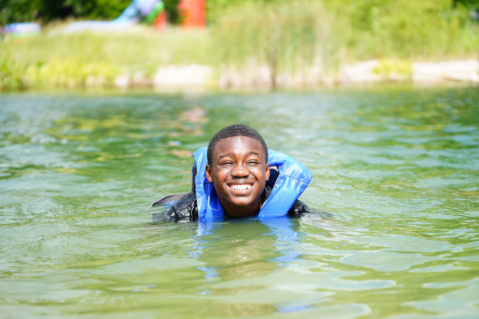 Young man smiling while swimming in a greenish lake with trees in the background.