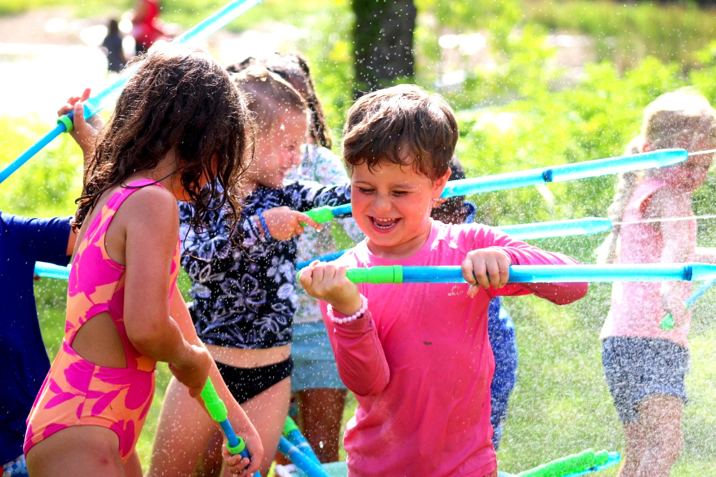 Children playing with water hoses outdoors on a sunny day, laughing and splashing water.