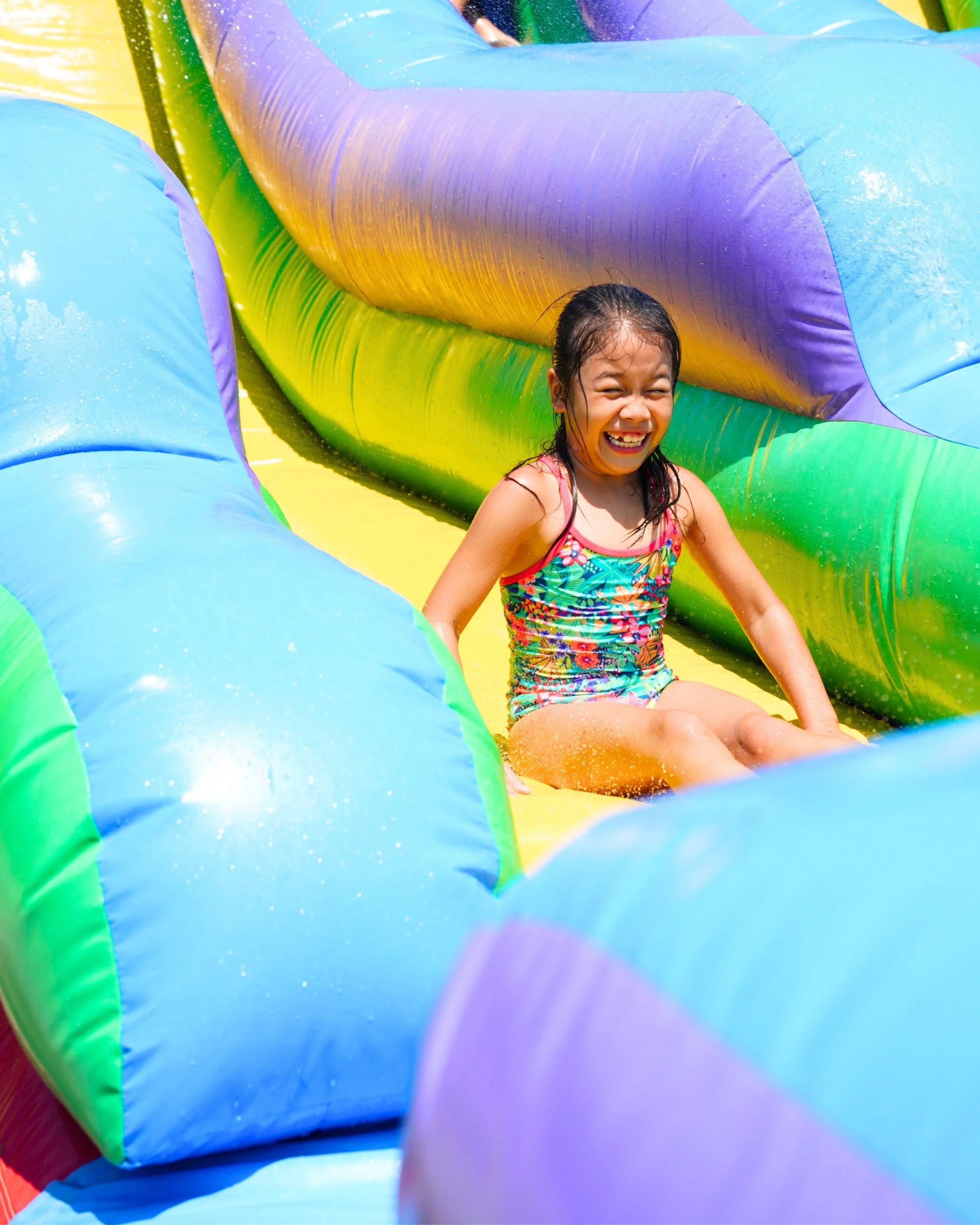 A girl with wet hair and a colorful swimsuit smiling while sitting at the bottom of an inflatable water slide or play area with bright yellow, green, blue, purple, and pink colors.