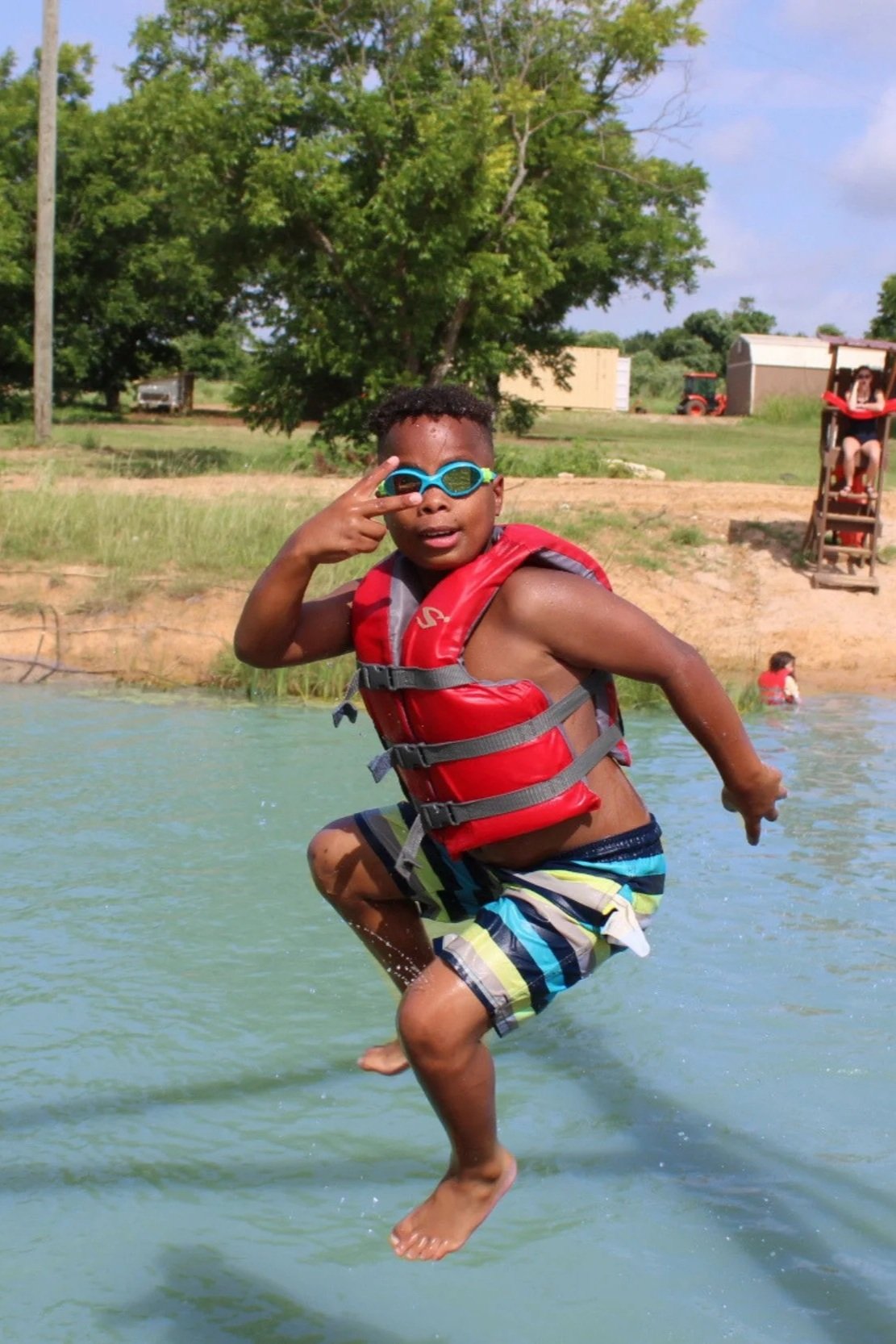 Young boy jumping in water wearing a red life jacket and goggles, making a peace sign with his right hand, with trees and a playground in the background.
