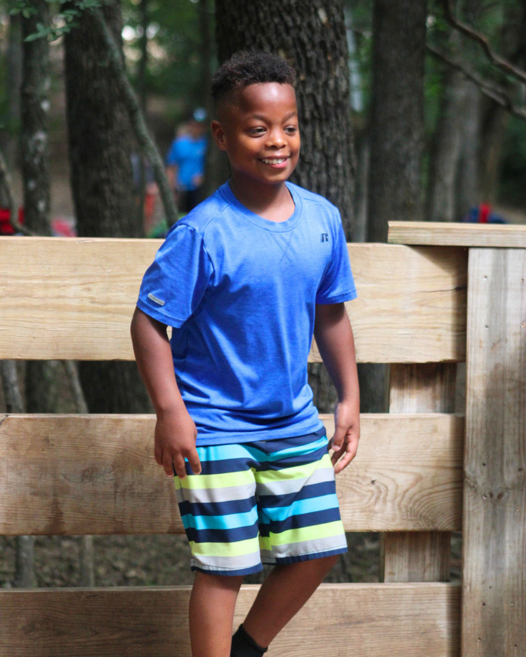 A young boy in a blue shirt and striped shorts standing on a wooden bridge in a forest.