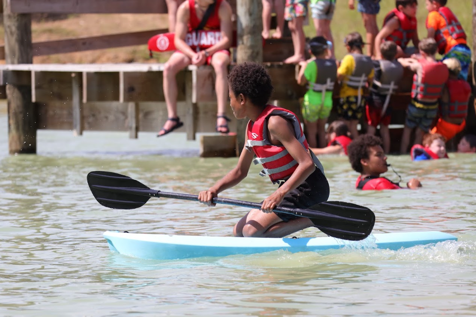 A boy in a red life jacket paddling a paddleboard in a lake with children swimming and people sitting on a dock in the background.