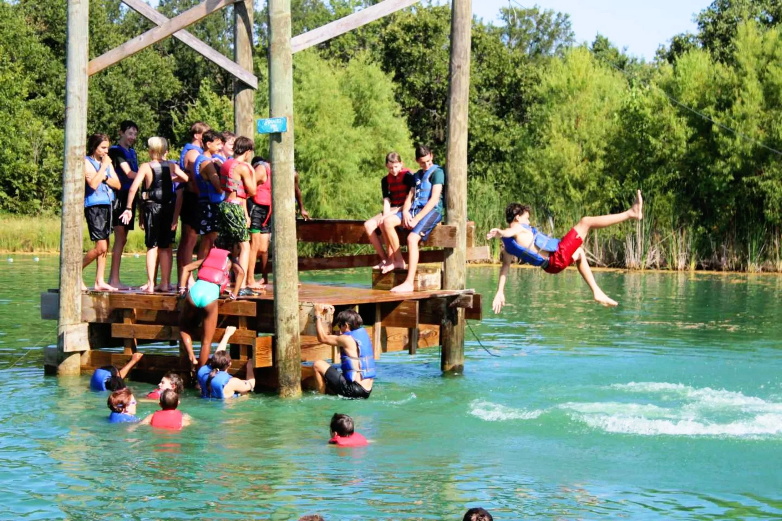 A group of children at a lake, some wearing life jackets, on a wooden dock with a boy jumping into the water, surrounded by green trees on a sunny day.