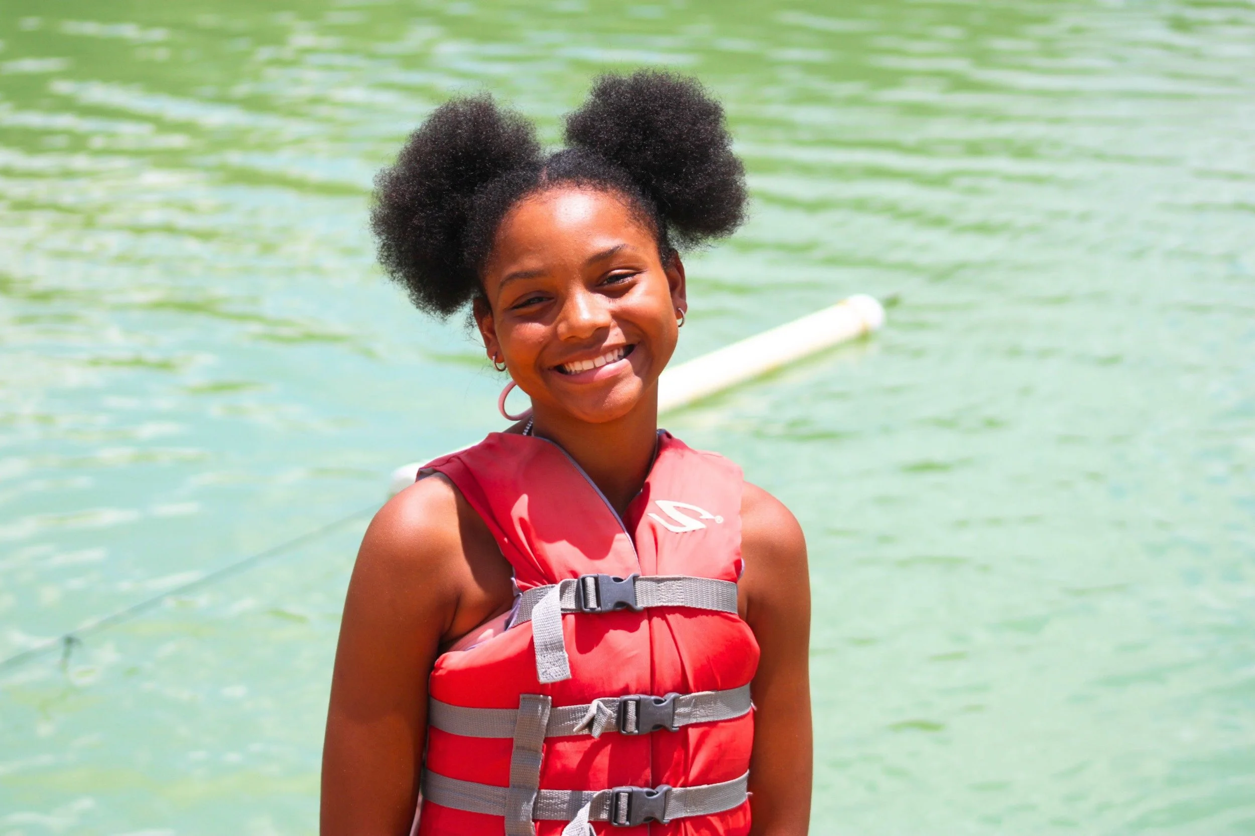 A young woman with dark curly hair styled in two puffs, smiling while standing by a body of water, wearing a red life jacket.