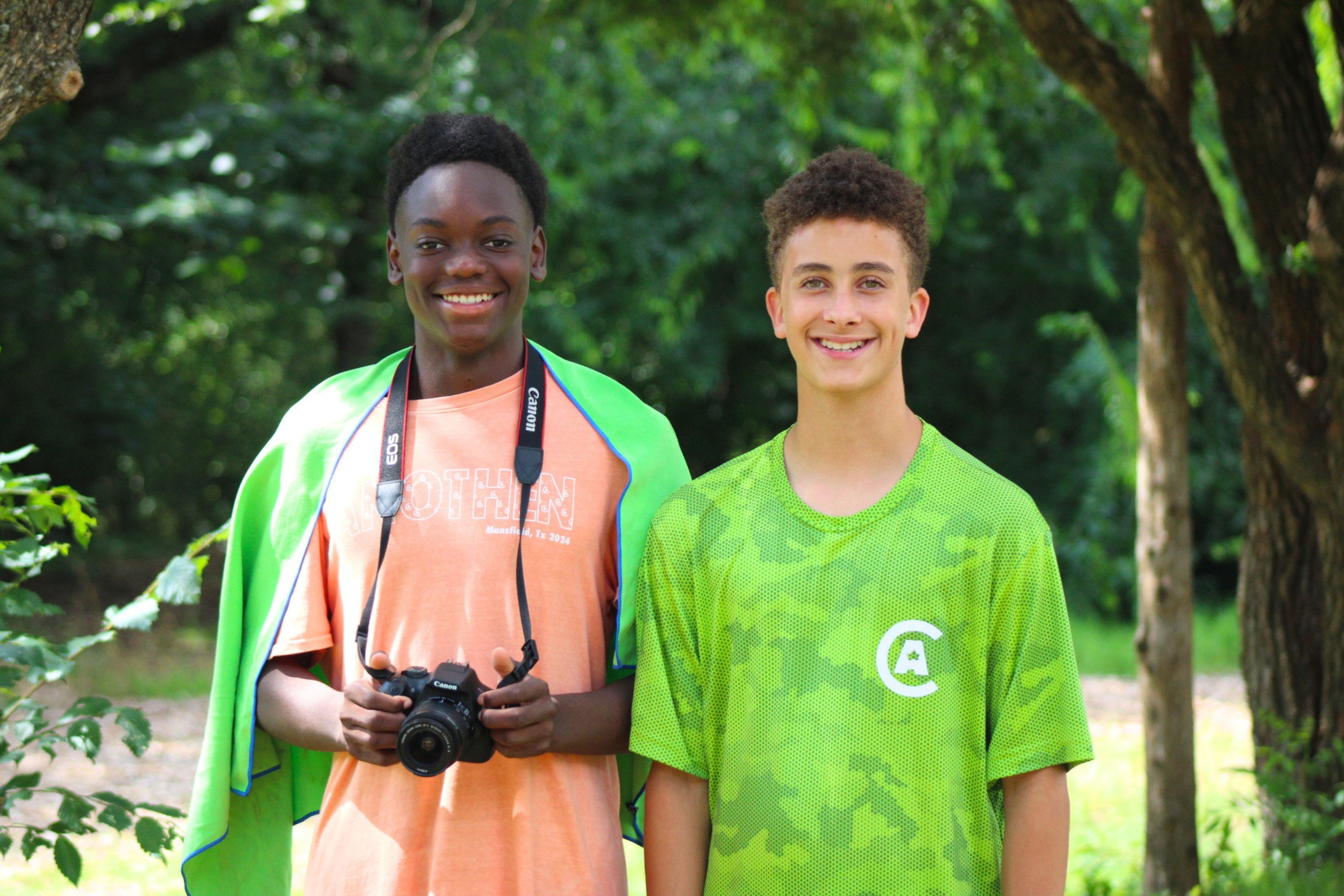 Two smiling boys standing outdoors in a park with green trees in the background. The boy on the left is holding a camera and wearing a light orange T-shirt with a green jacket draped over his shoulders. The boy on the right is wearing a bright green sports jersey.
