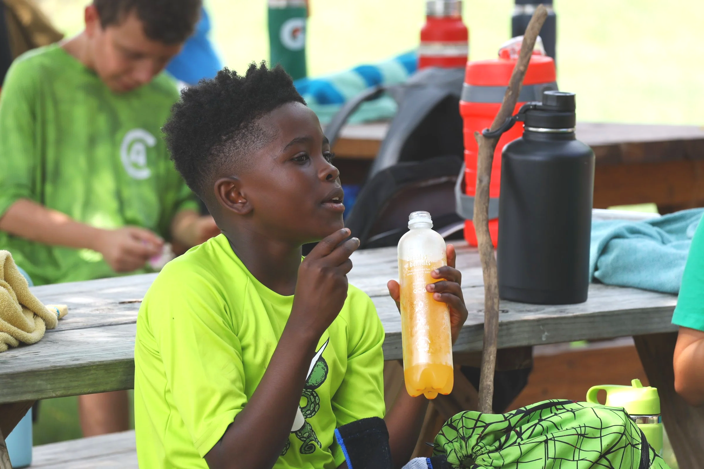 A young boy in a bright yellow shirt sitting at a picnic table outdoors, holding a yellow water bottle, with various outdoor gear and beverages on the table. Other children are visible in the background.