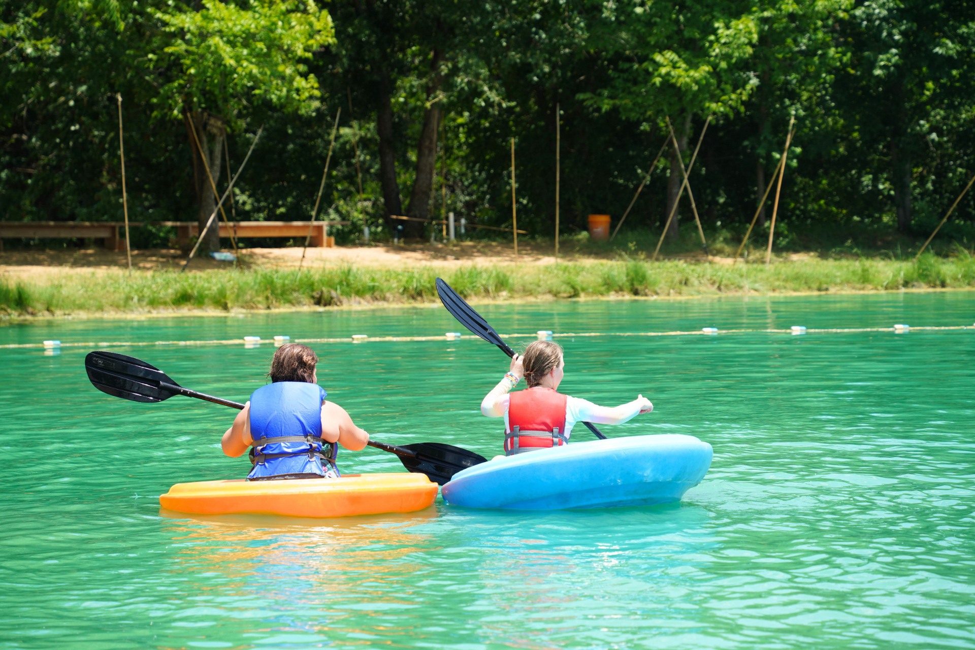 Two people kayaking on a lake with green water, wearing life jackets, with a wooded shoreline and a wooden deck in the background.