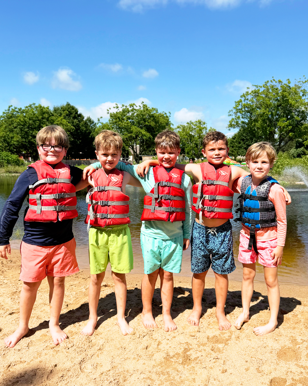 Five young boys standing side by side on a sandy beach, wearing life jackets and summer shorts, with arms around each other's shoulders, by a lake with trees and blue sky in the background.