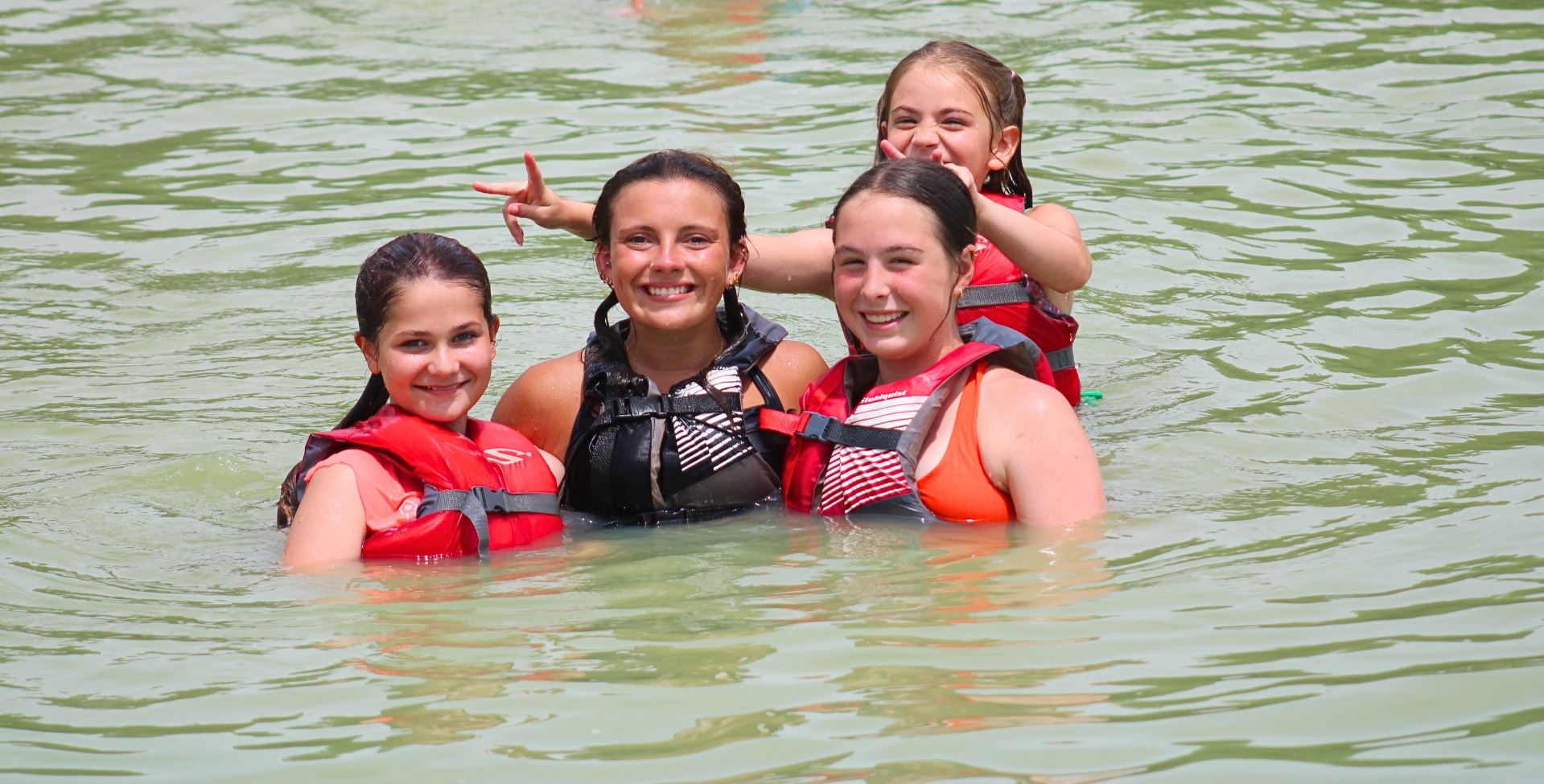 Five young girls in life jackets enjoying a swim in a river or lake, smiling and making playful gestures.