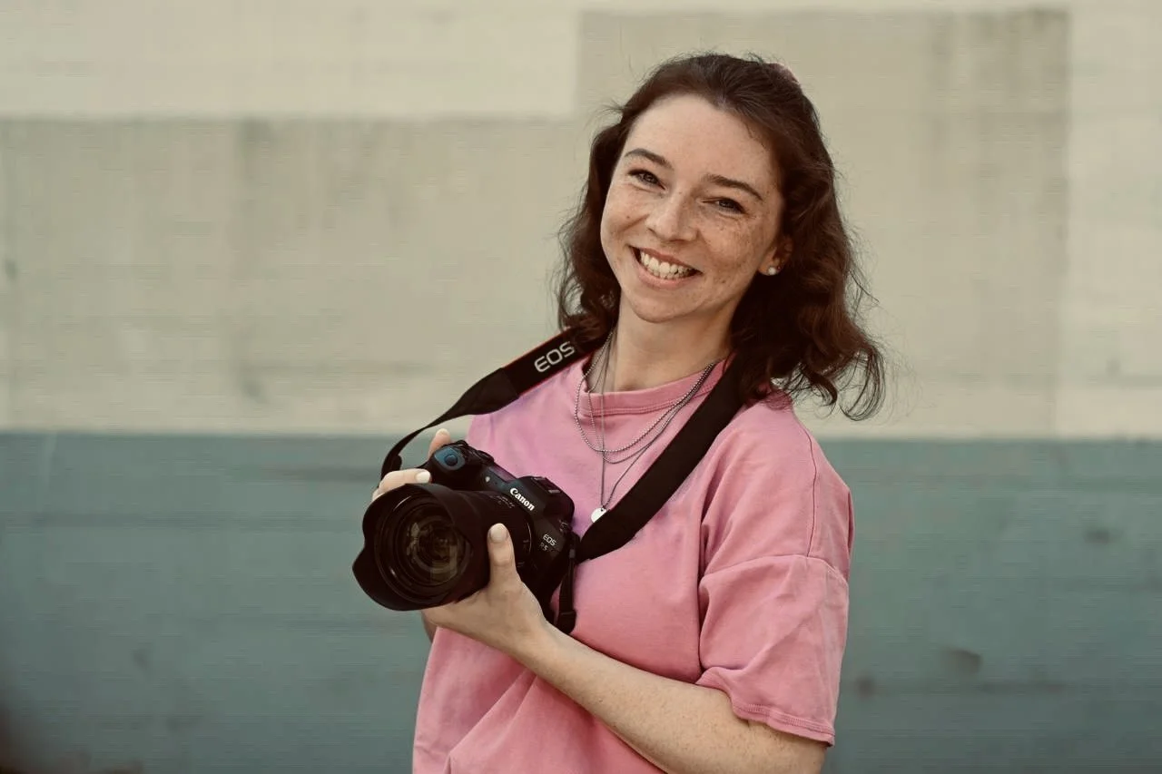 Eine junge Frau mit lockigem braunem Haar, trägt ein rosa T-Shirt und hält eine Digitalkamera in den Händen. Sie lächelt freundlich. Canon R5, Fotografin, Michelle Rehberg.