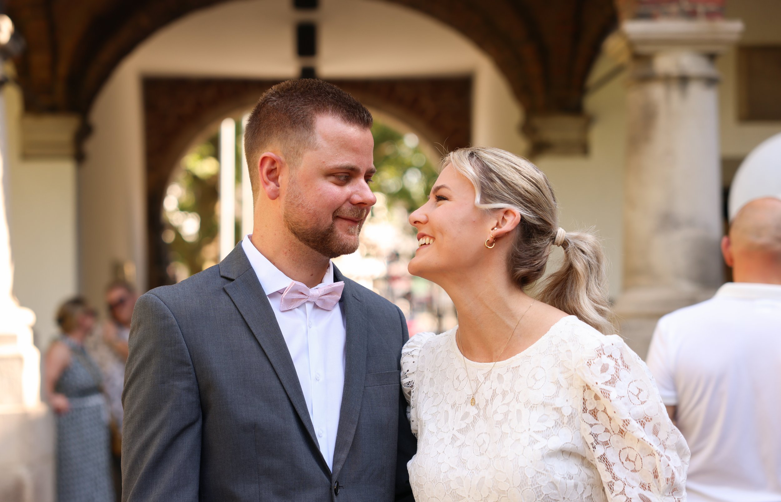 Ein verliebtes Paar bei einer Hochzeit, das sich liebevoll in die Augen schaut, vor einer historischen Kulisse mit Bögen und Säulen. Standesamt, Trauung.