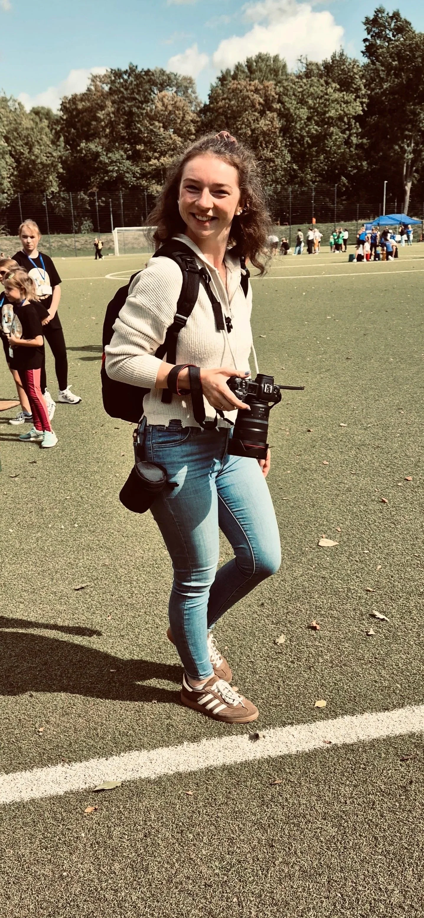 Junge Frau mit Kamera auf einem Fußballfeld, umgeben von Kindern und Erwachsenen, im Freien bei sonnigem Wetter.