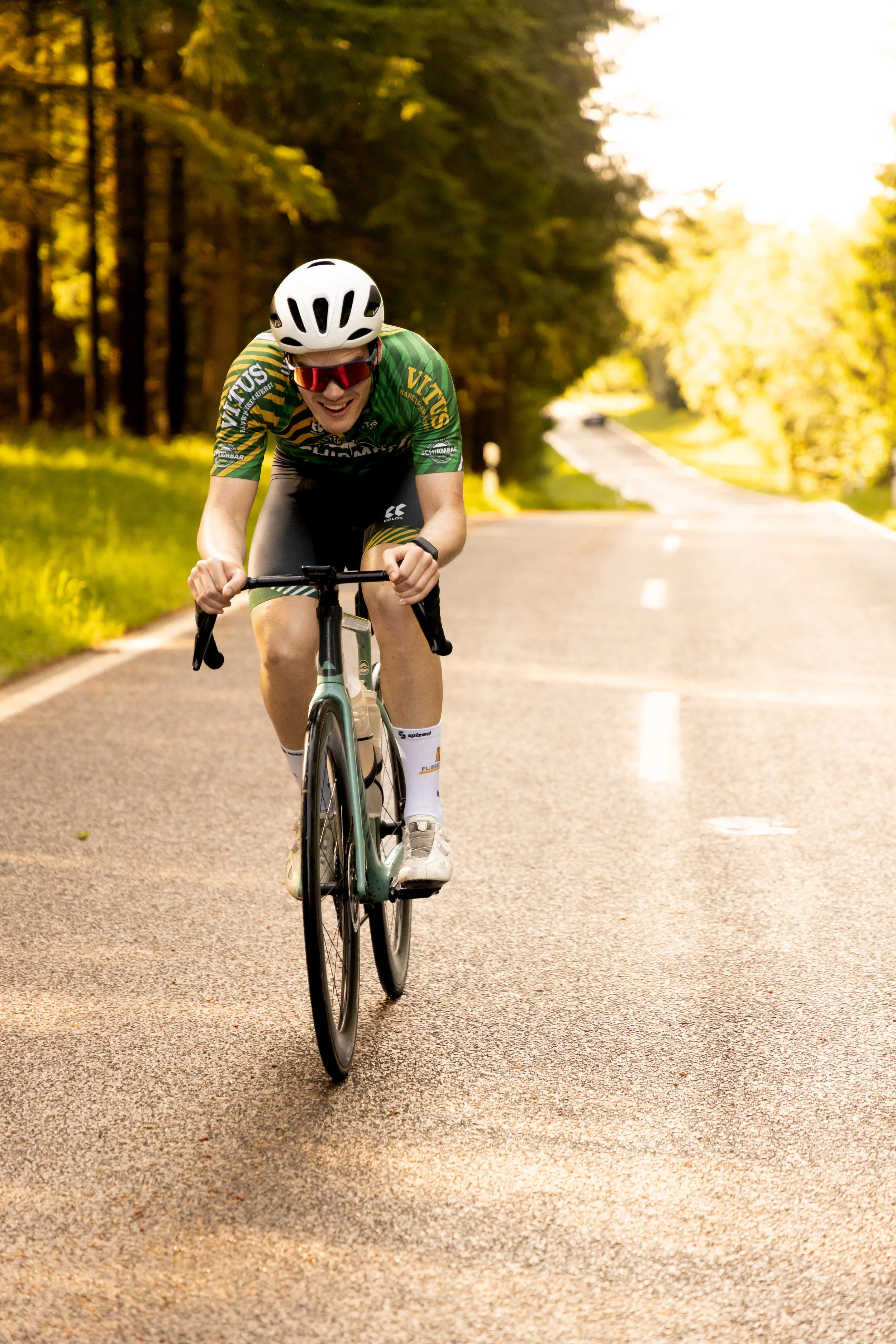 Ein Radfahrer in Sonnenbrille und Helm fährt auf einer ruhigen Landstraße durch einen bewaldeten Bereich bei Sonnenuntergang. Sport, Sport-Shooting, Sport-Fotografie, Rennrad.