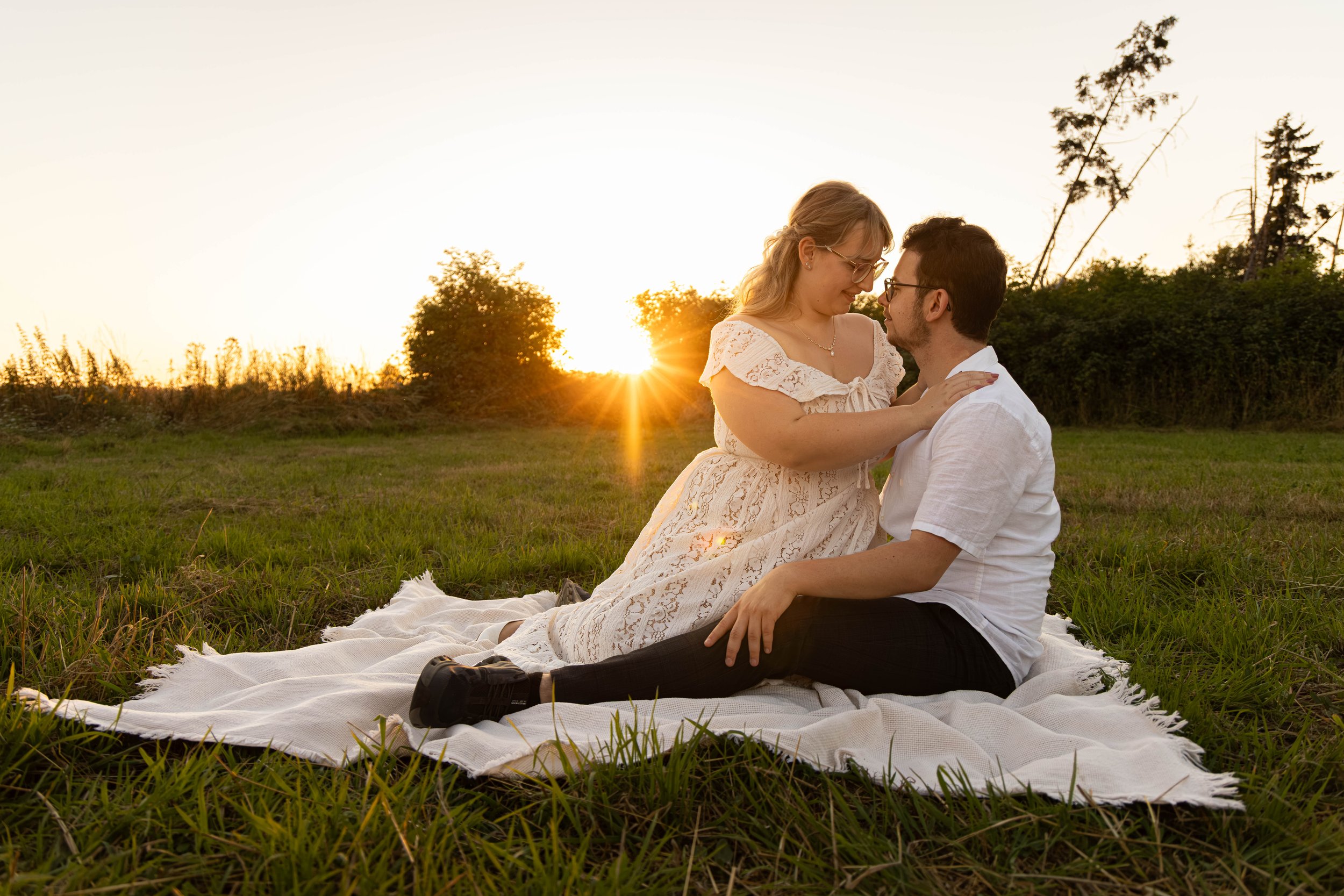 Ein Paar sitzt auf einer Decke im Gras, umgeben von Bäumen, bei Sonnenuntergang, miteinander verbunden, lächelnd, zärtlich. Sunset-Shooting, Sunset Shooting, Golden Hour Shooting, Golden Hour-Shooting.