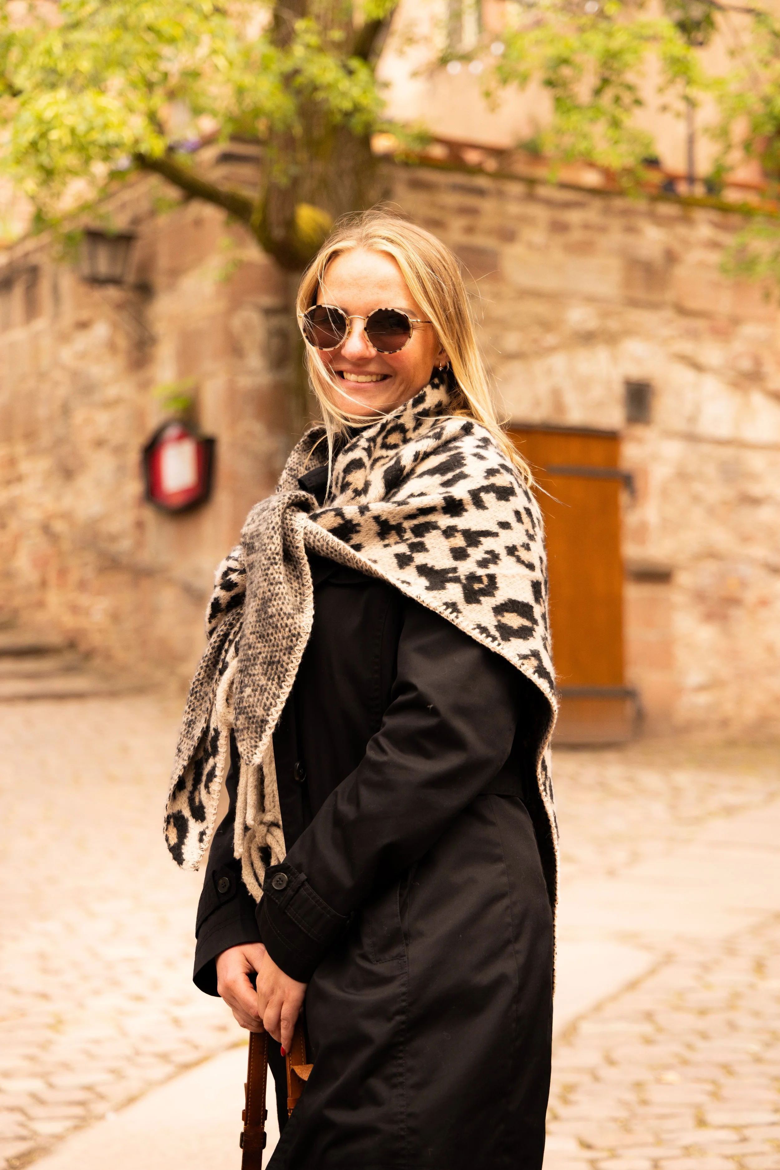 Eine lachende blonde Frau trägt eine Sonnenbrille, einen schwarzen Mantel und einen Leopardenmuster-Schal, während sie draußen auf einem Kopfsteinpflasterweg vor einer Steinmauer mit grünem Baum steht. Porträt, Porträt-Shooting.