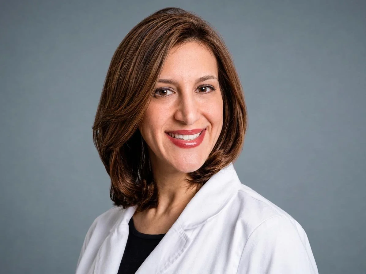 A woman with shoulder-length brown hair smiling at the camera, wearing a white lab coat over a black top against a plain gray background.