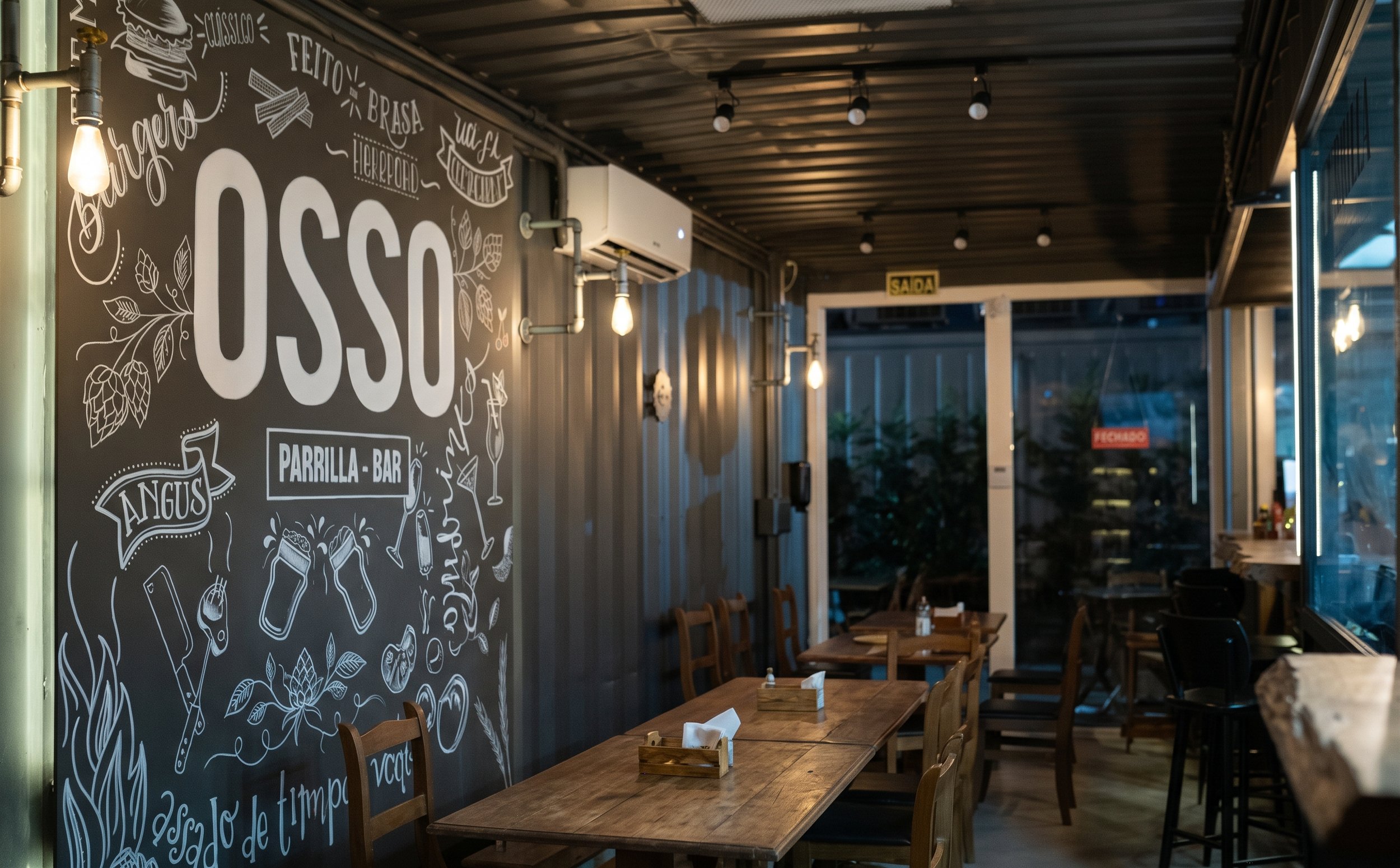 Salão do Osso Parrilla Bar em Canoas, com mesas de madeira e parede de lousa desenhada