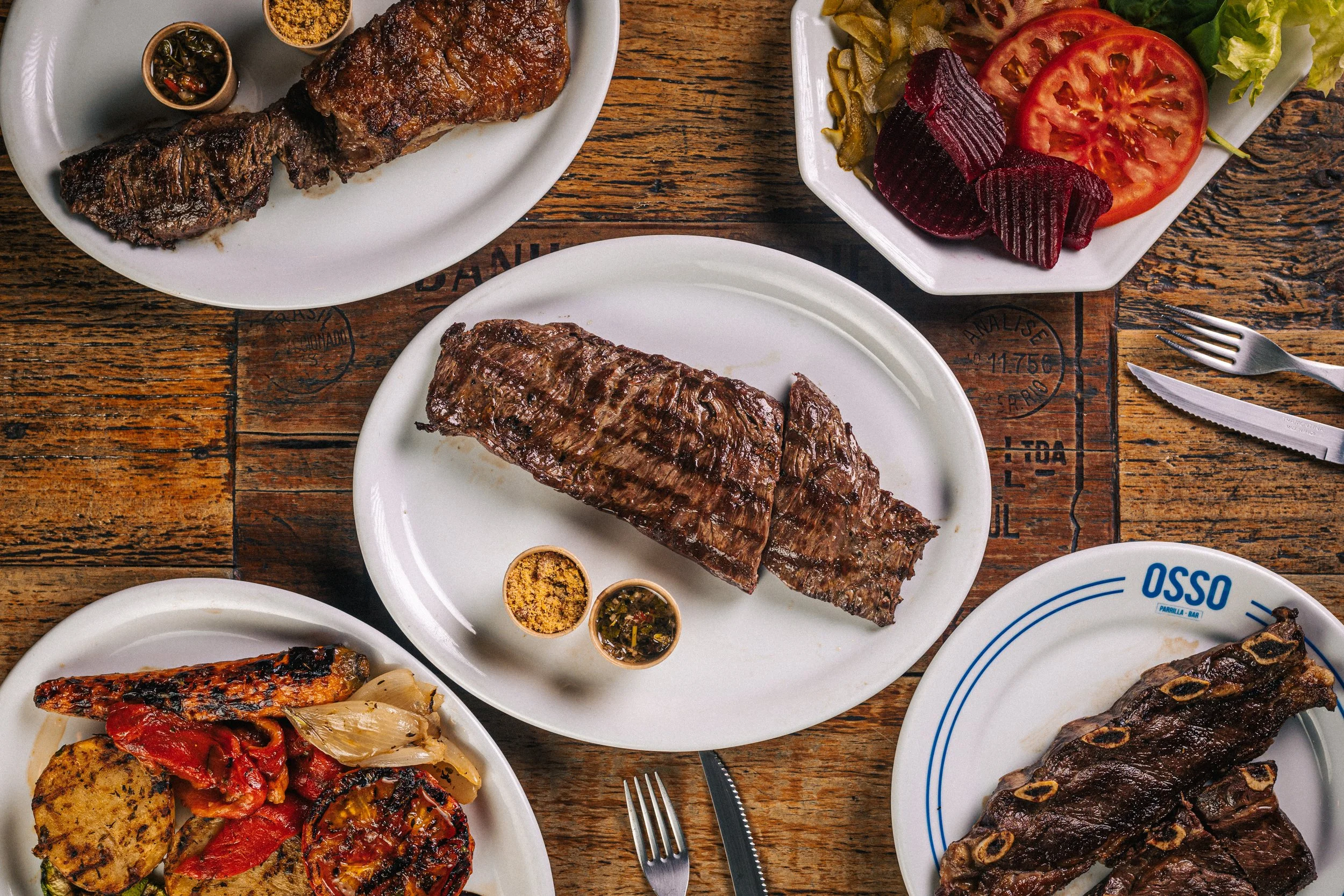 Mesa com cortes de carne grelhados na parrilla e acompanhamentos em pratos do Osso