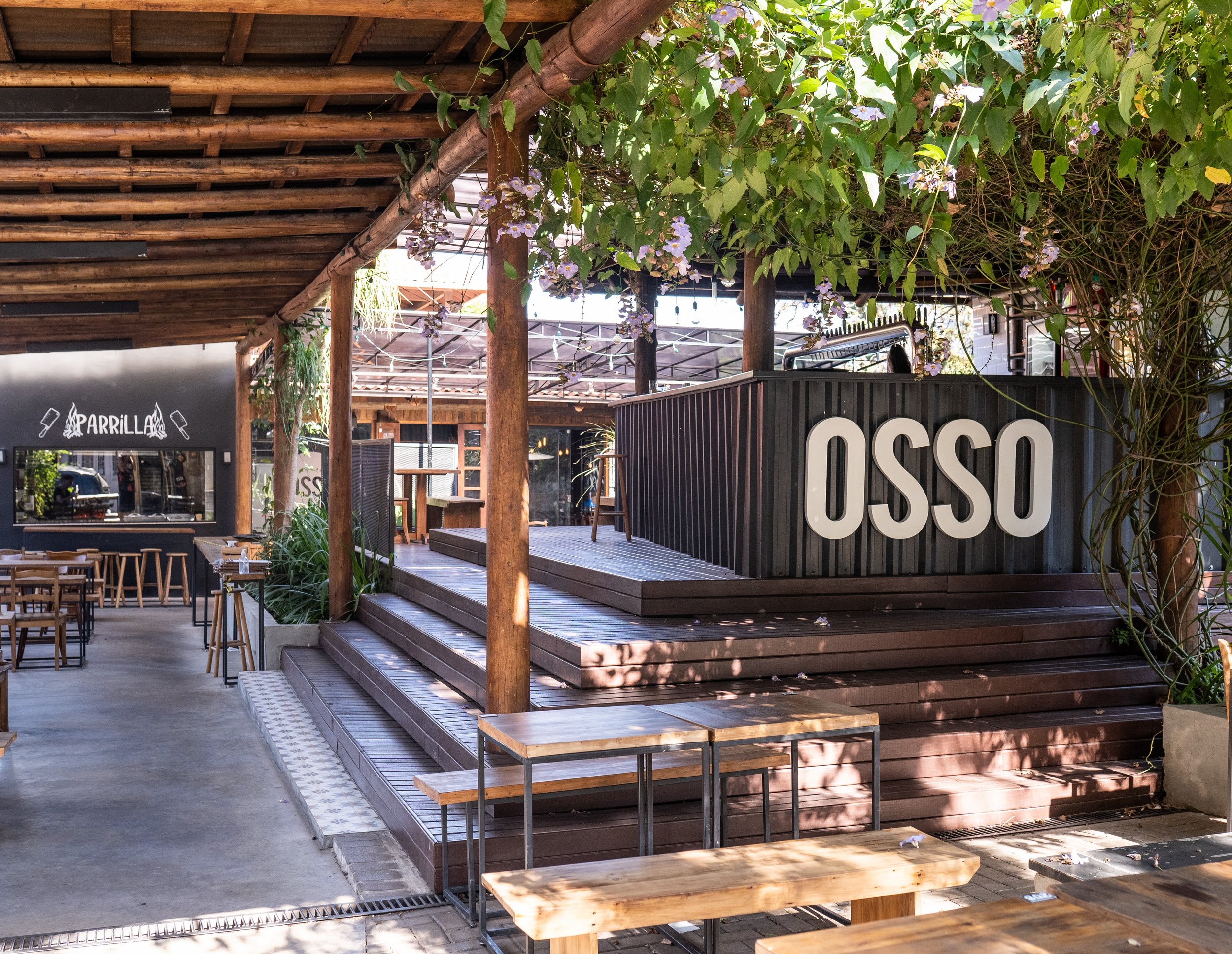 Pátio do Osso Parrilla Bar no Bom Fim, com degraus e o muro preto "OSSO" sob a cobertura de madeira