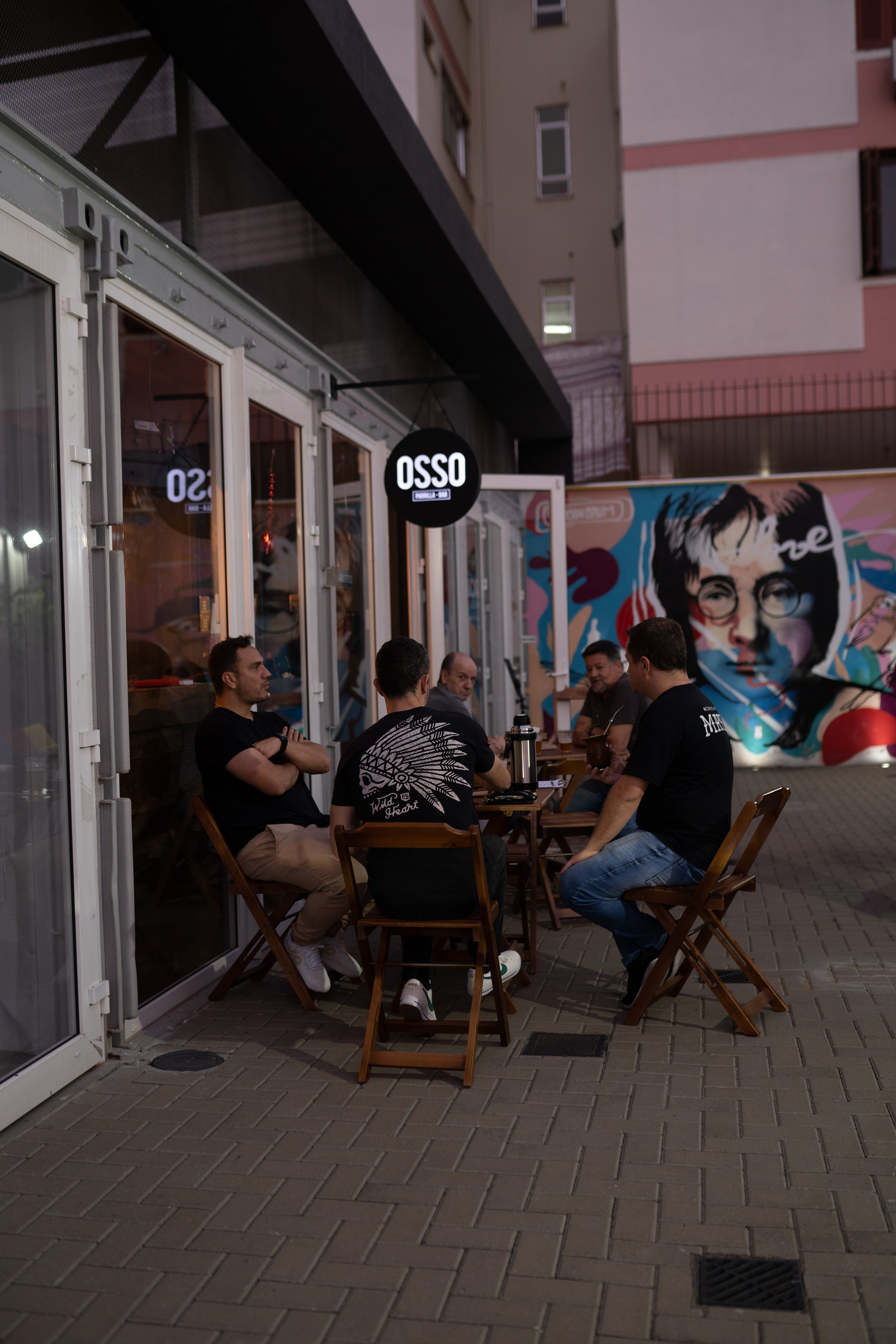 Clientes sentados em mesas na calçada do Osso Parrilla Bar em Canoas, ao lado de um mural