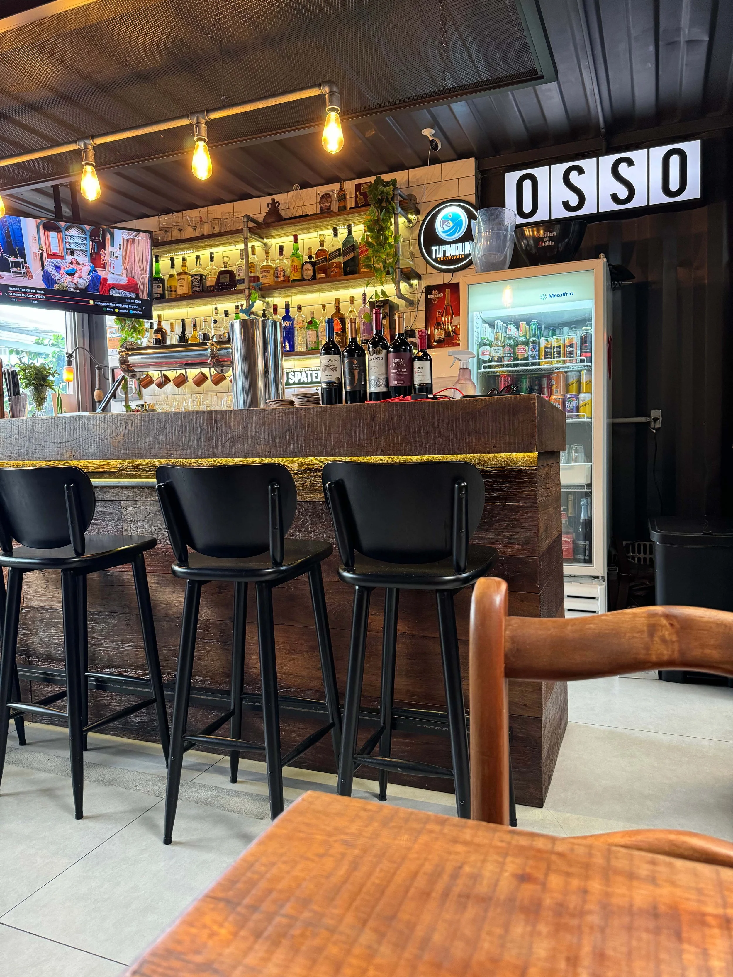 Balcão do bar do Osso Parrilla Bar em Canoas, com banquetas pretas, prateleiras de bebidas e letreiro OSSO