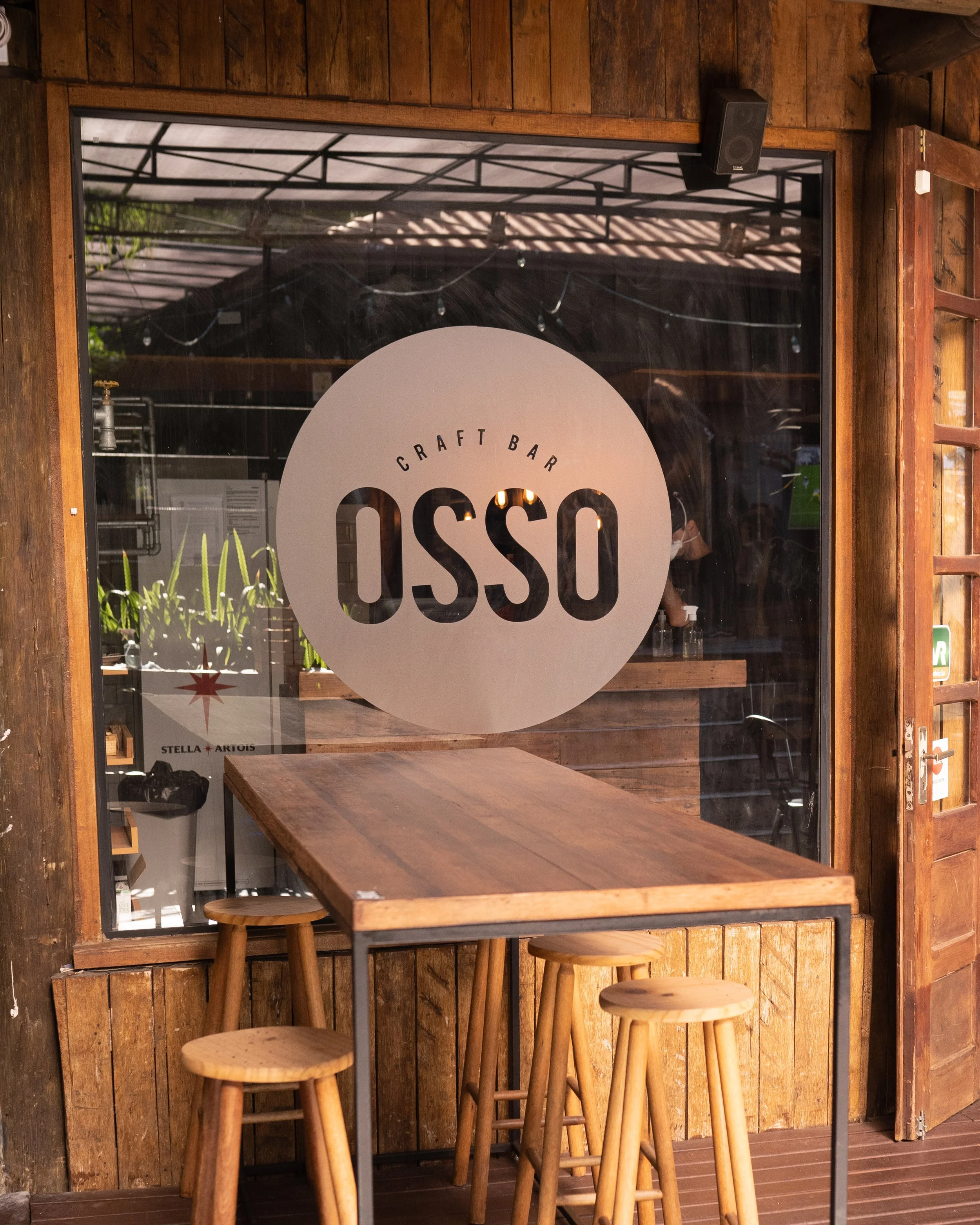 Fachada de vidro do Osso Parrilla Bar no Bom Fim, com o letreiro "Craft Bar OSSO" e banquetas de madeira
