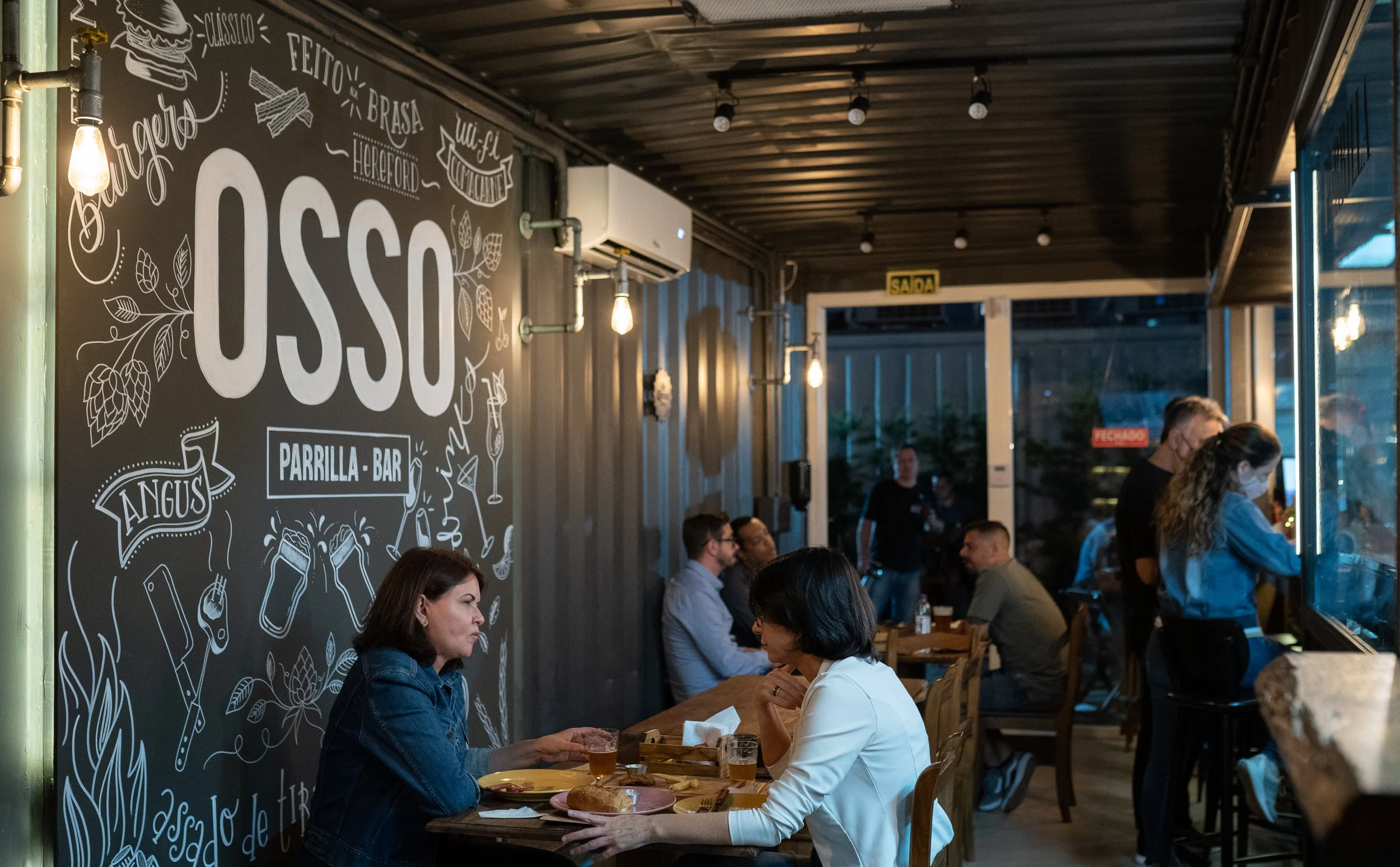 Clientes à mesa no salão do Osso Parrilla Bar em Canoas, com a lousa "OSSO Parrilla Bar" ao fundo