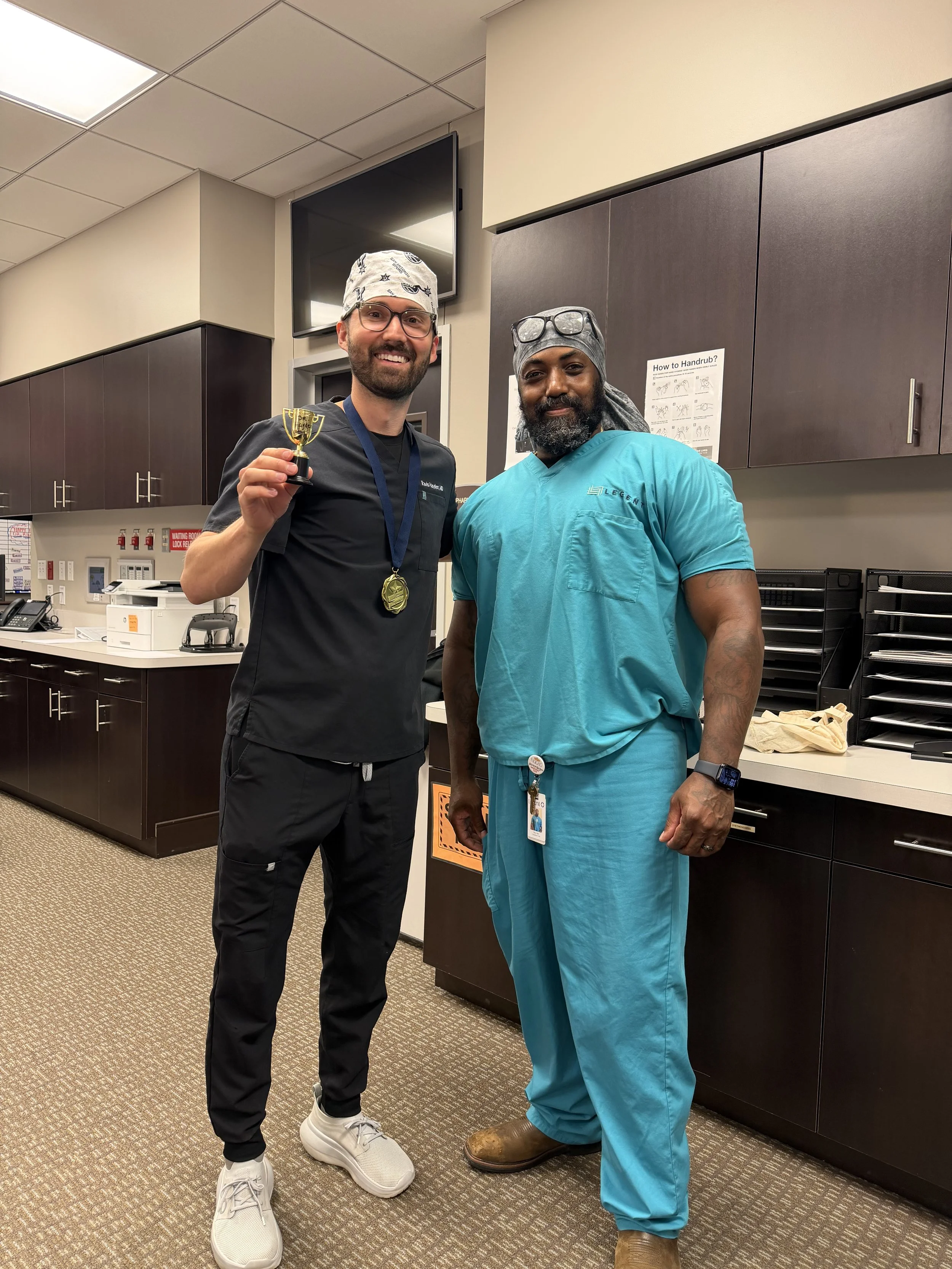 Two men standing in a medical office, smiling at the camera. One man is holding a small gold trophy and wearing black scrubs, glasses, and a bandana. The other man is wearing blue scrubs, boots, and glasses on his head.