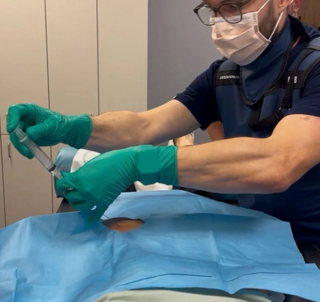 A healthcare professional, wearing green gloves, a face mask, glasses, and scrubs, prepares a syringe while performing a medical procedure on a patient lying on an operating table covered with blue surgical drapes.