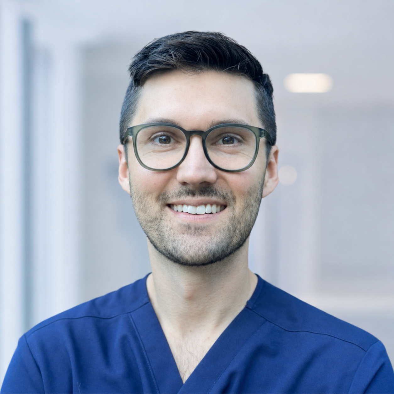A smiling man in navy medical scrubs and glasses, with short dark hair, standing in a modern indoor setting.