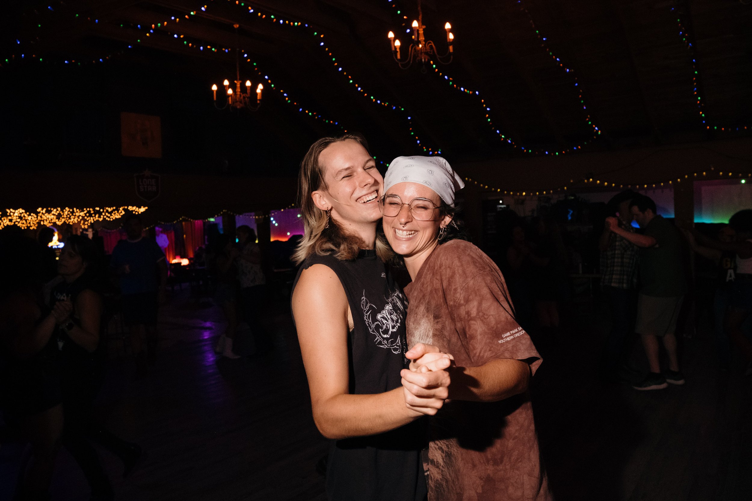 Two women are dancing and smiling at a party with festive colorful string lights and chandeliers hanging from the ceiling.