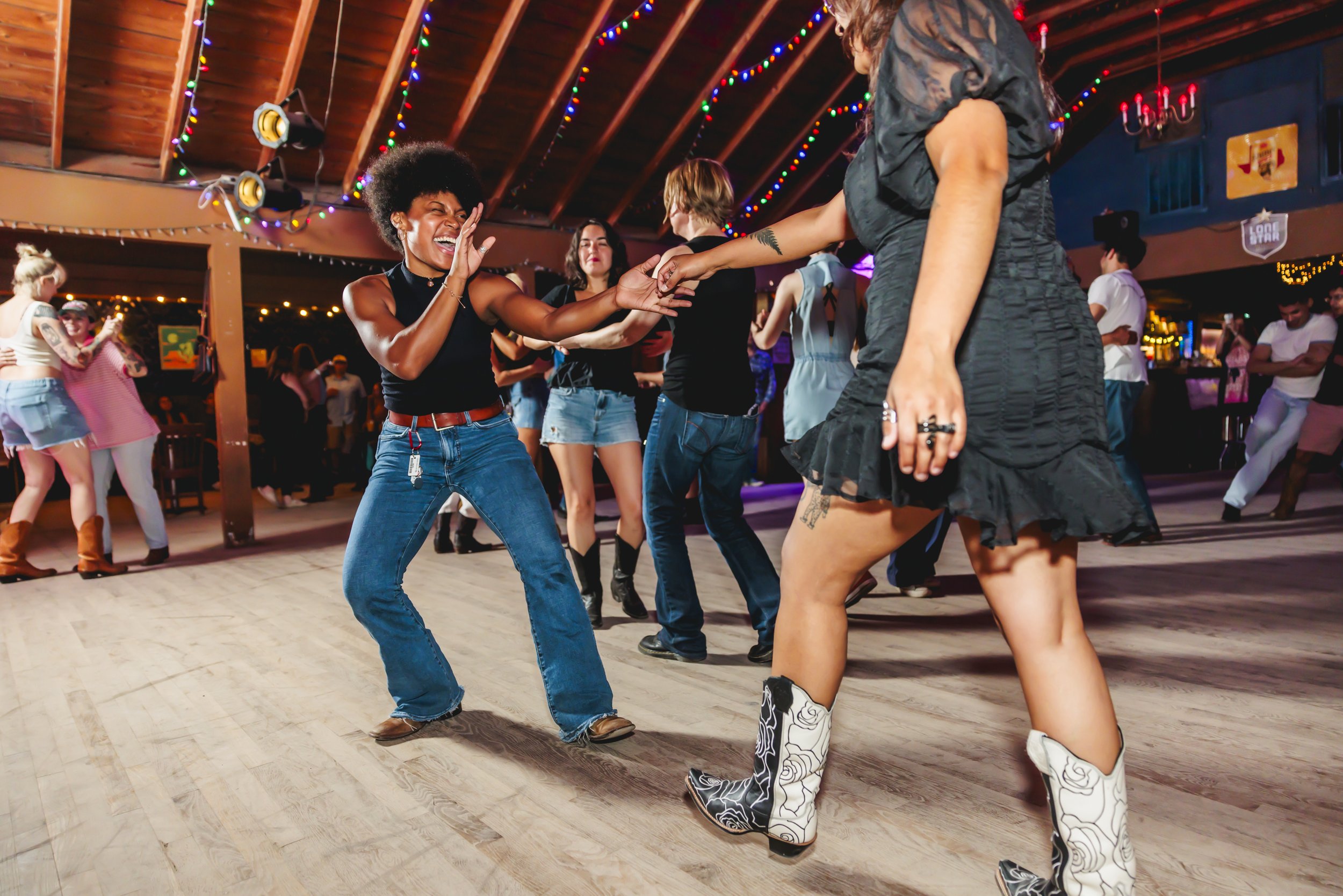 People at a country dance or two step event, dancing on a wooden floor decorated with colorful string lights against a rustic wood ceiling. Bar, South Austin