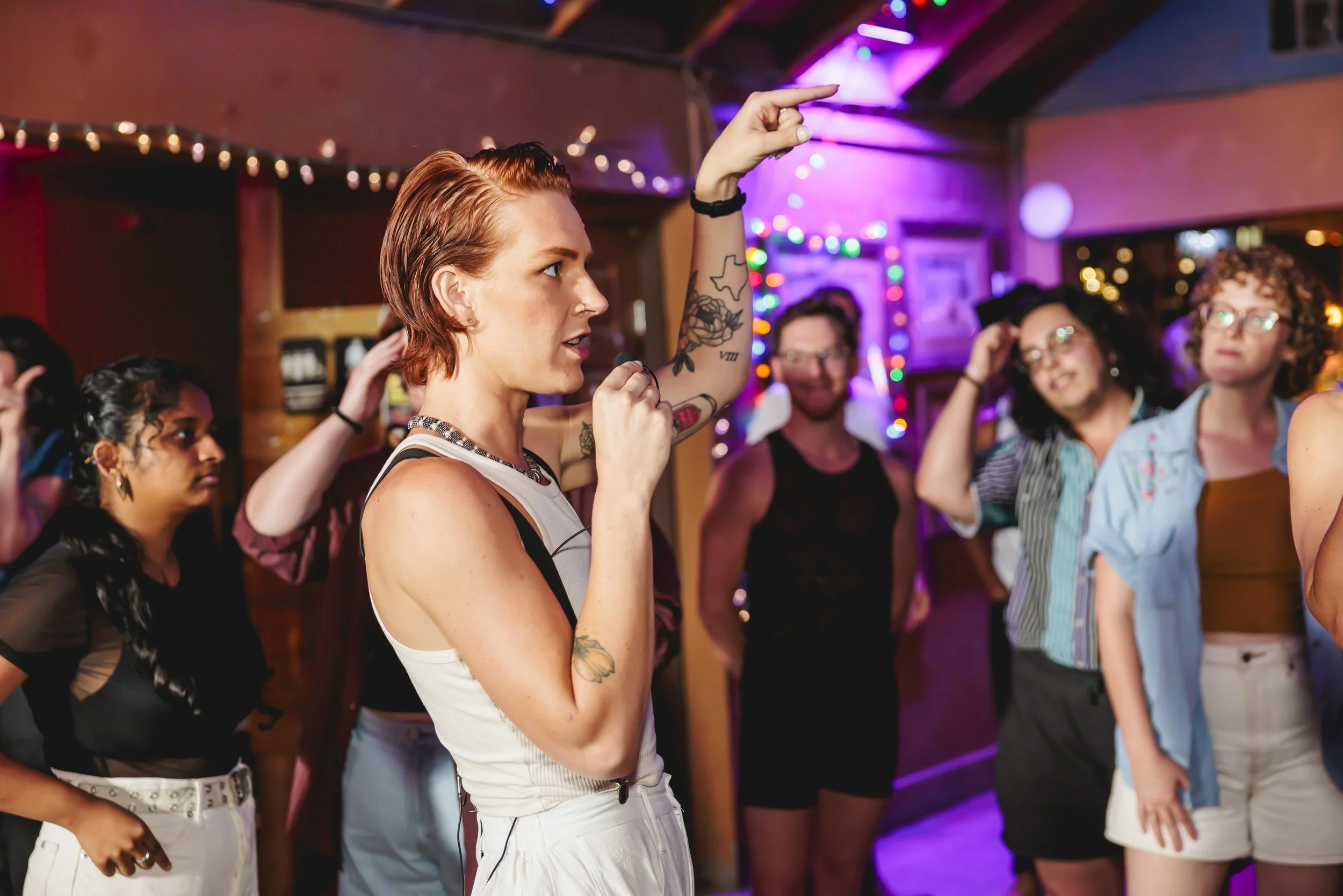 A woman with short red hair, tattoos on her arm, and wearing a sleeveless top, points upward with her right hand while teaching a group two step lesson at Sagebrush in South Austin, Texas. A crowd around her listens and learns.