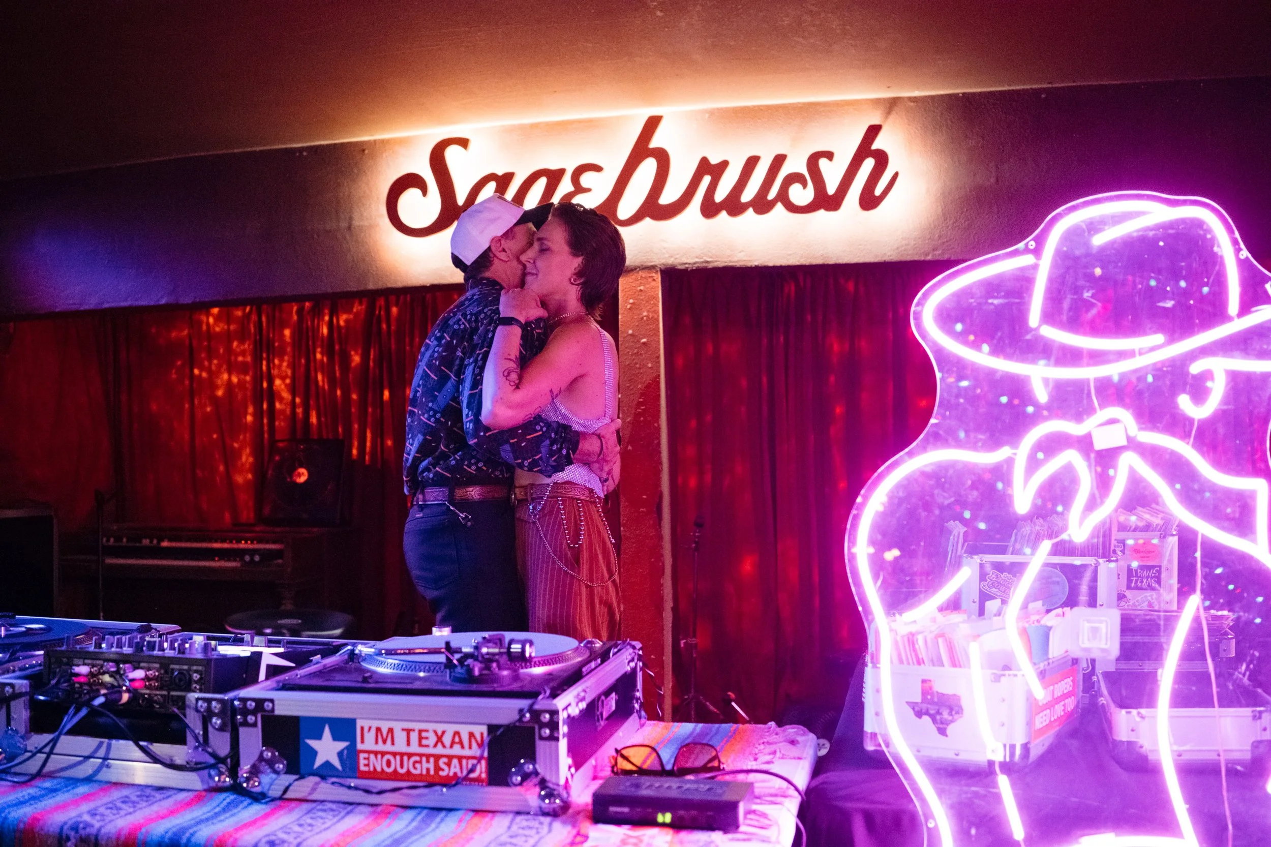 A queer couple is dancing closely together in a dimly lit bar with neon signage and red curtains in the background. There is a DJ setup with turntables and equipment in the foreground. The woman has tattoos, is smiling, and the man is wearing a cap.