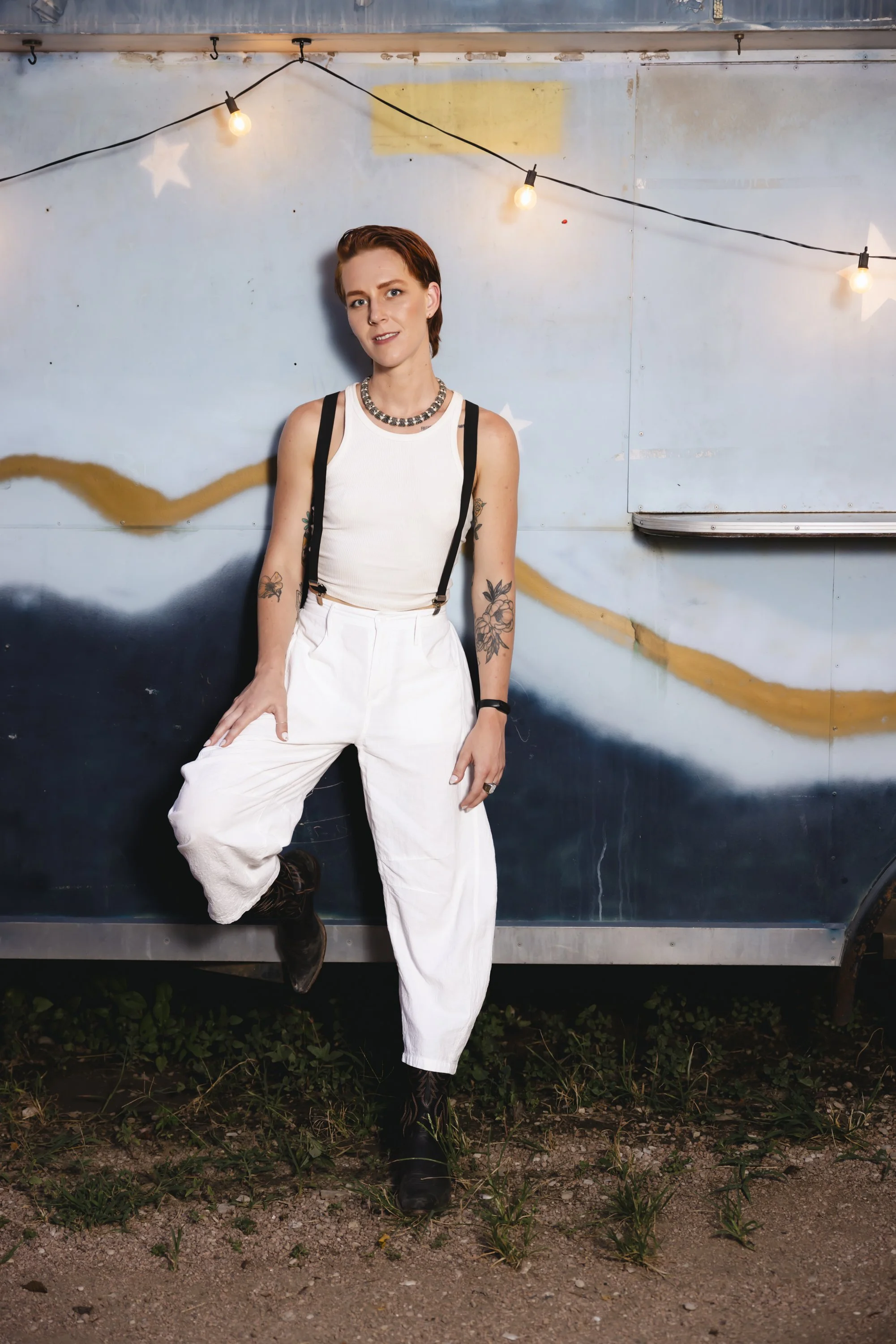 A person with short red hair and tattoos on their arms, wearing a white tank top, suspenders, white pants, and dark boots, leaning against a blue wall with string lights hanging above, and holding one leg up. The ground has dirt and small plants.