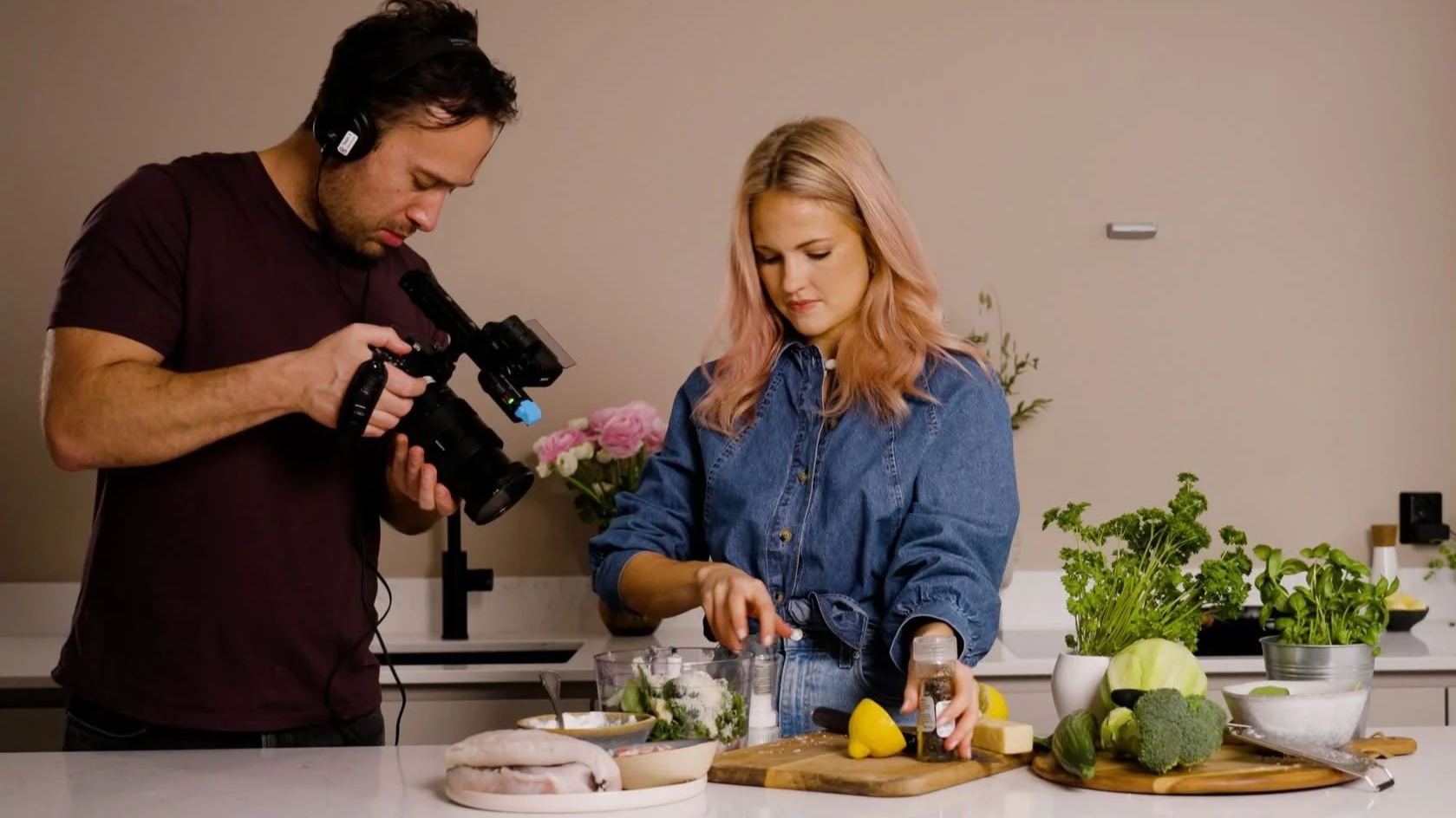 A woman with pink hair in a denim jacket prepares vegetables and ingredients on a kitchen counter, while a man films her with a camera. The counter has lettuce, broccoli, and other greens, and there are flowers and a faucet in the background.