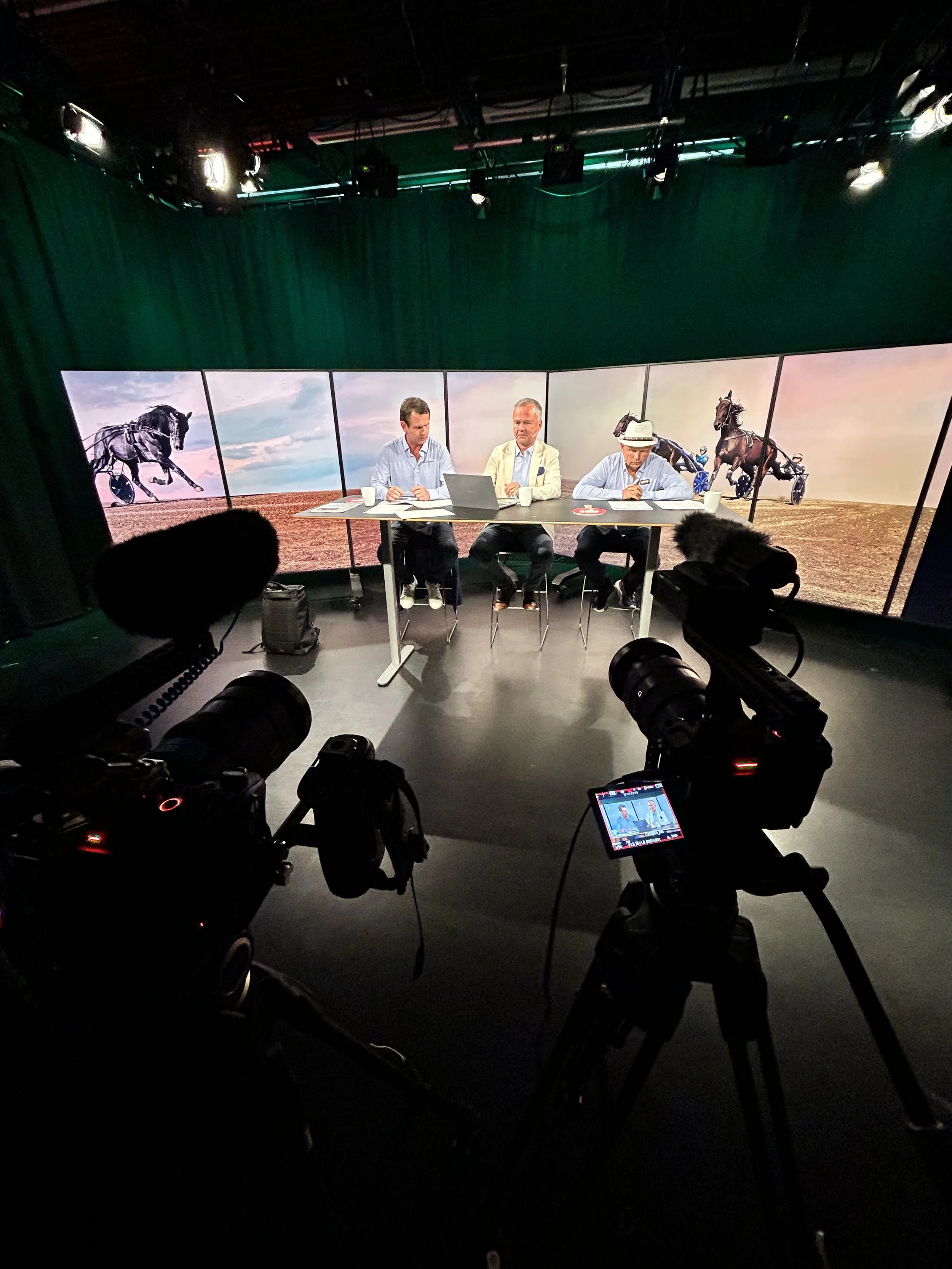 Studio recording setup with three men sitting at a table in front of a large screen displaying horse racing images. Cameras and equipment are visible in the foreground.