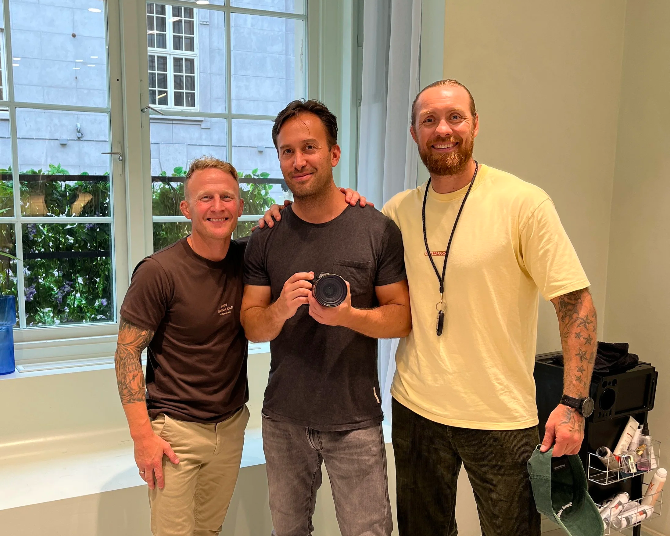 Three men standing inside a room with a large window. The man on the left has short, blond hair, tattoos on his right arm, and is wearing a brown shirt and beige pants. The man in the middle has dark, wavy hair, and is wearing a dark gray t-shirt, holding a camera. The man on the right has a beard, long hair tied back, and is wearing a yellow t-shirt with tattoos on his left arm, and a lanyard around his neck. They are smiling for the camera.